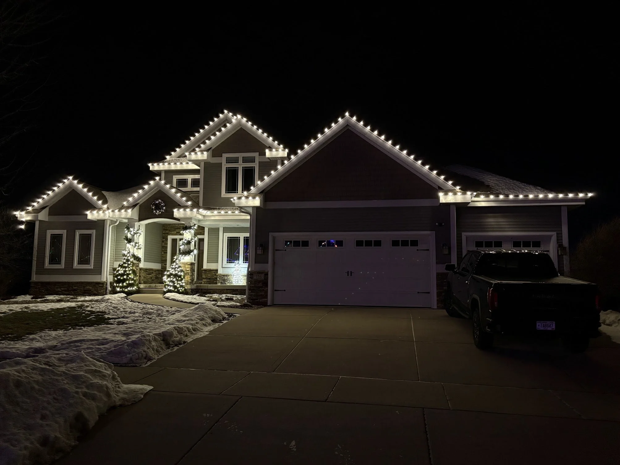 A house decorated with Christmas lights, with snow on the ground and trees at the side, at night.