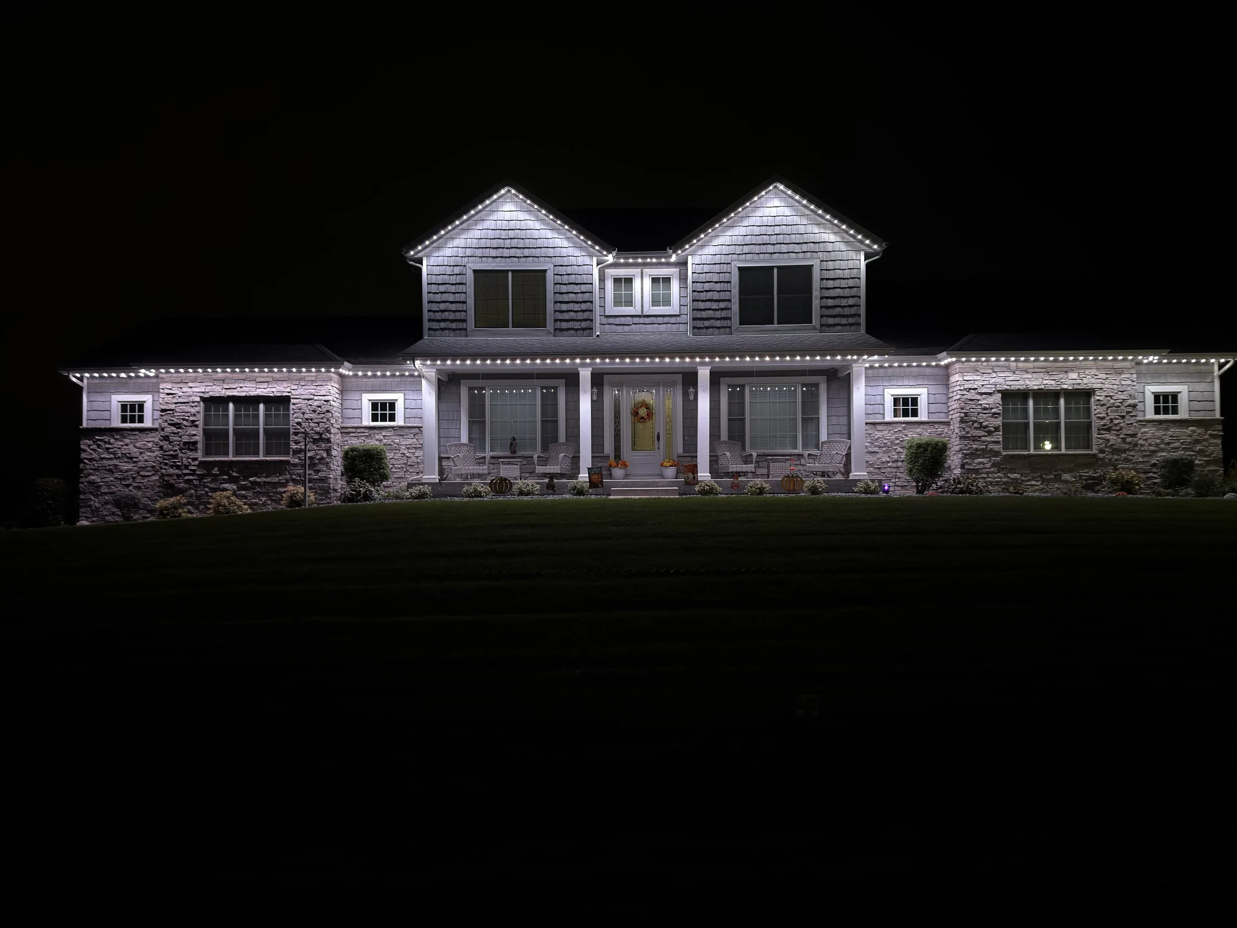 A house decorated with white permanent LED lights outlining the roof, windows, and porch at night. The house has stone and siding exterior with a small front yard.
