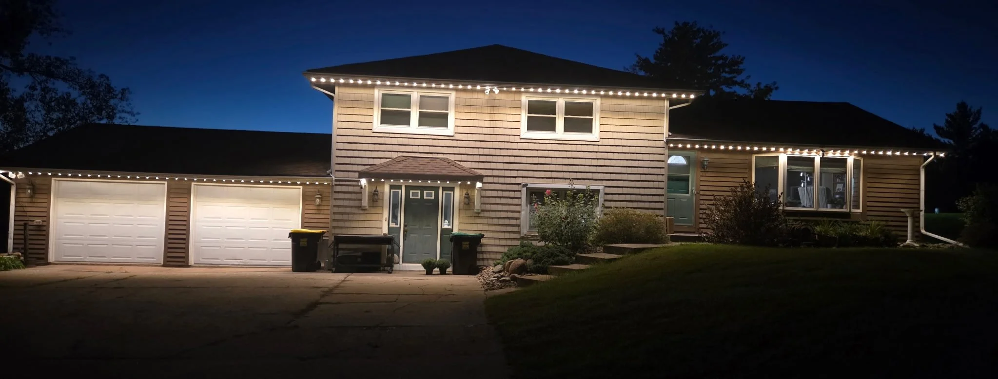 A two-story house at night decorated with permanent lighting along the roofline, featuring a three-car garage, a front door, windows, and a garden with bushes and plants.