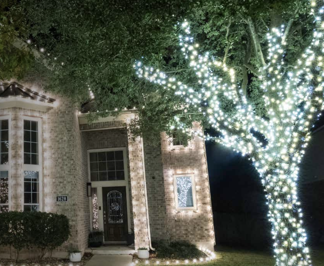 Nighttime scene of a house with brick exterior illuminated by string lights outlining the windows, driveway, and roof. A large tree in front is decorated with numerous white string lights, creating a festive display.