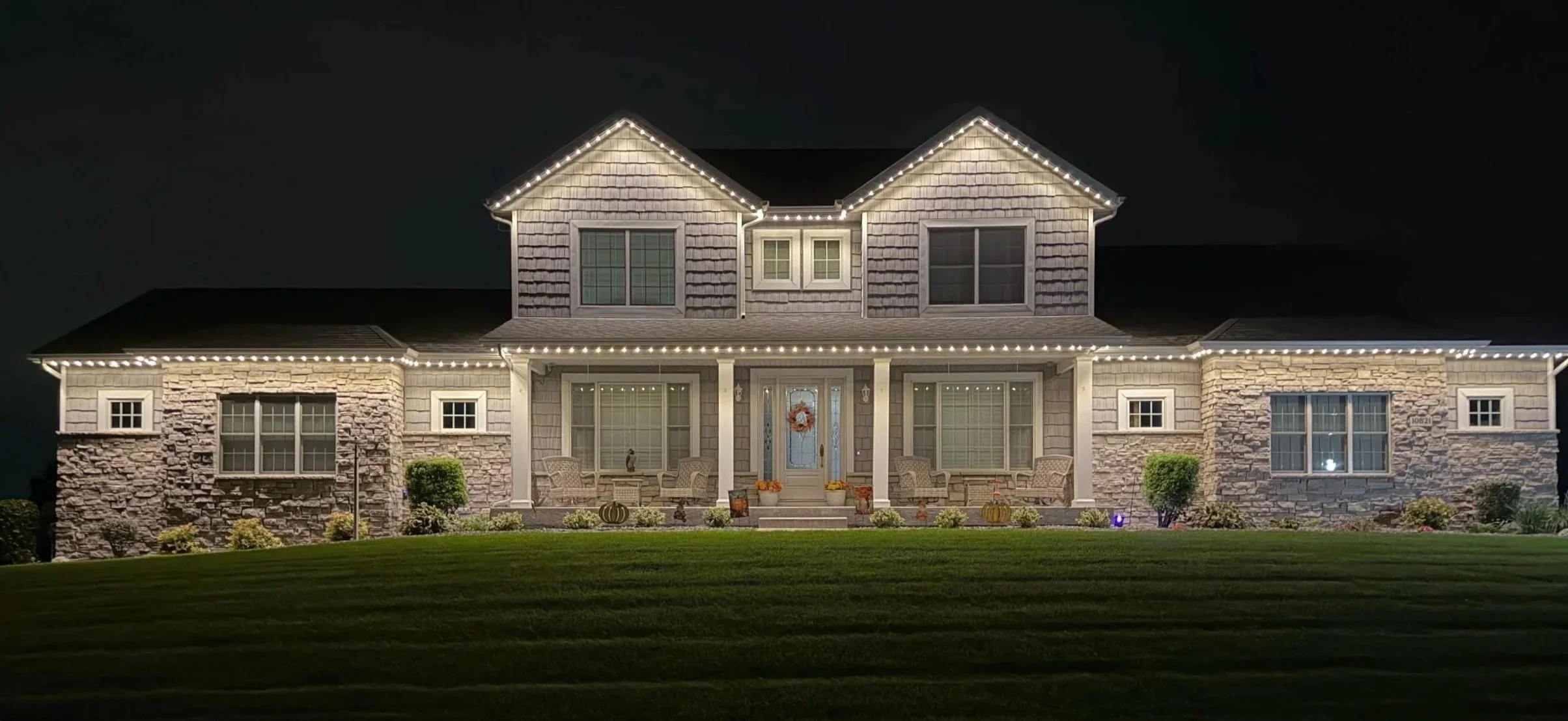 A large house decorated with permanent lights at night, featuring stone and siding exterior, multiple windows, a front porch with chairs, and a well-kept lawn.