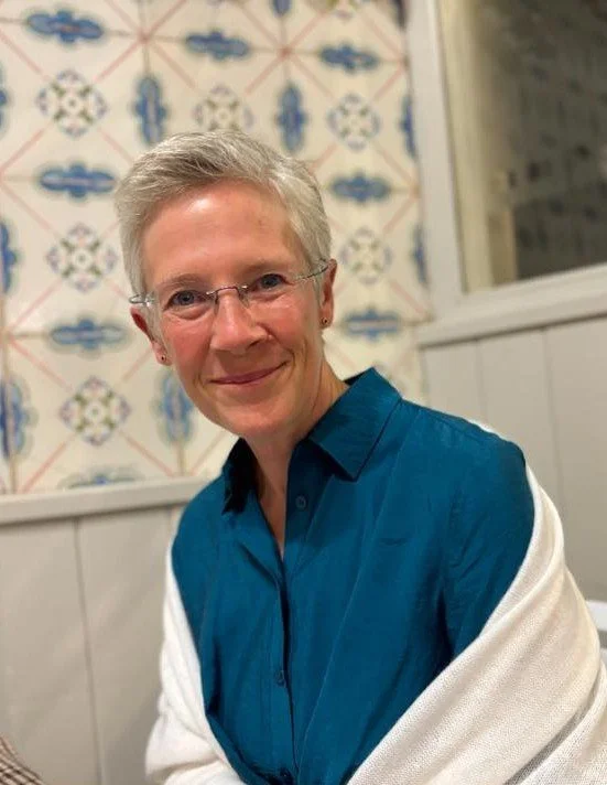A smiling woman with short gray hair and glasses sitting indoors in front of a tiled wall, wearing a blue shirt and a white sweater draped over her shoulders.