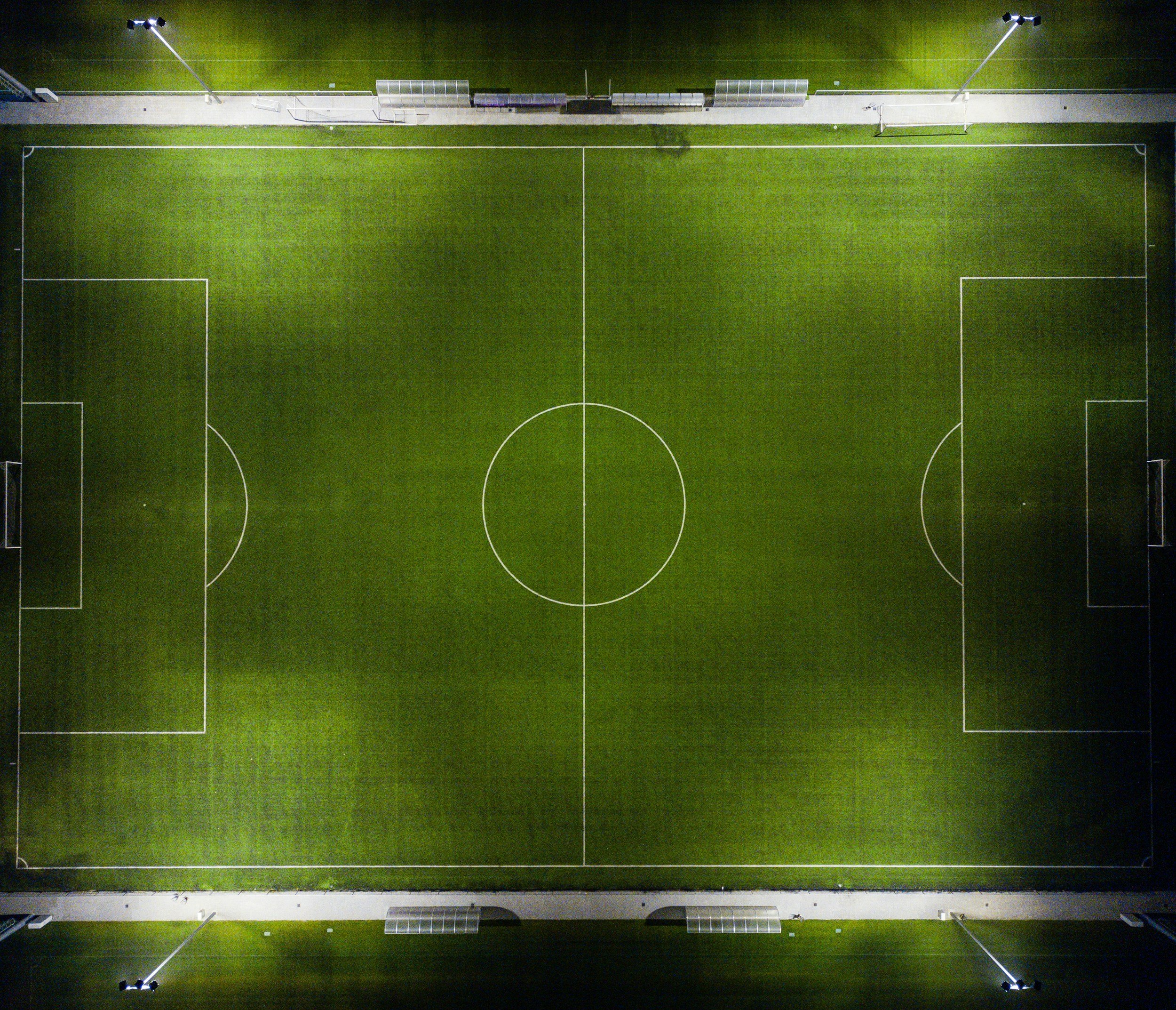 An aerial view of an empty illuminated soccer field at night, with marked white lines and a surrounding running track, illuminated by tall floodlights.