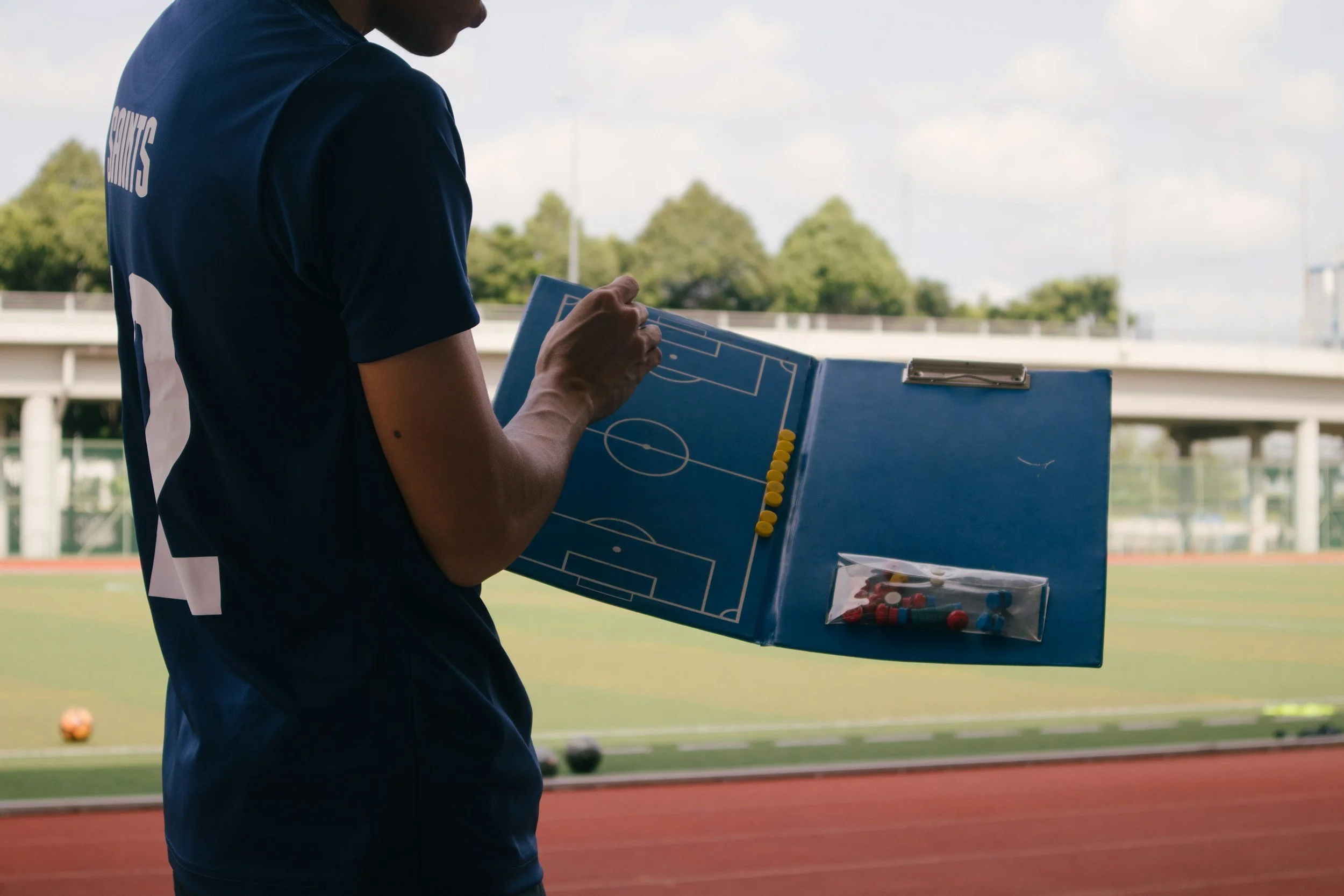 A person standing with their side to the camera on a sports field, holding a clipboard with a soccer field diagram, at a stadium.