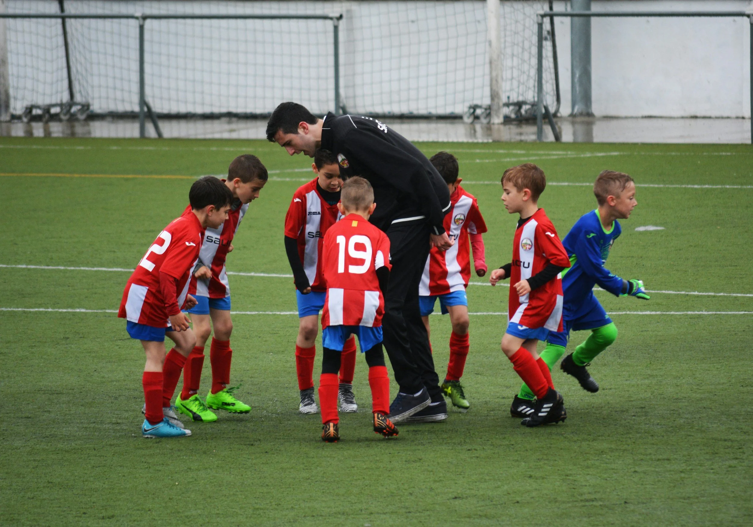 A group of young children wearing red and blue soccer uniforms and green cleats gather around a coach on a soccer field, listening attentively during a practice or game.