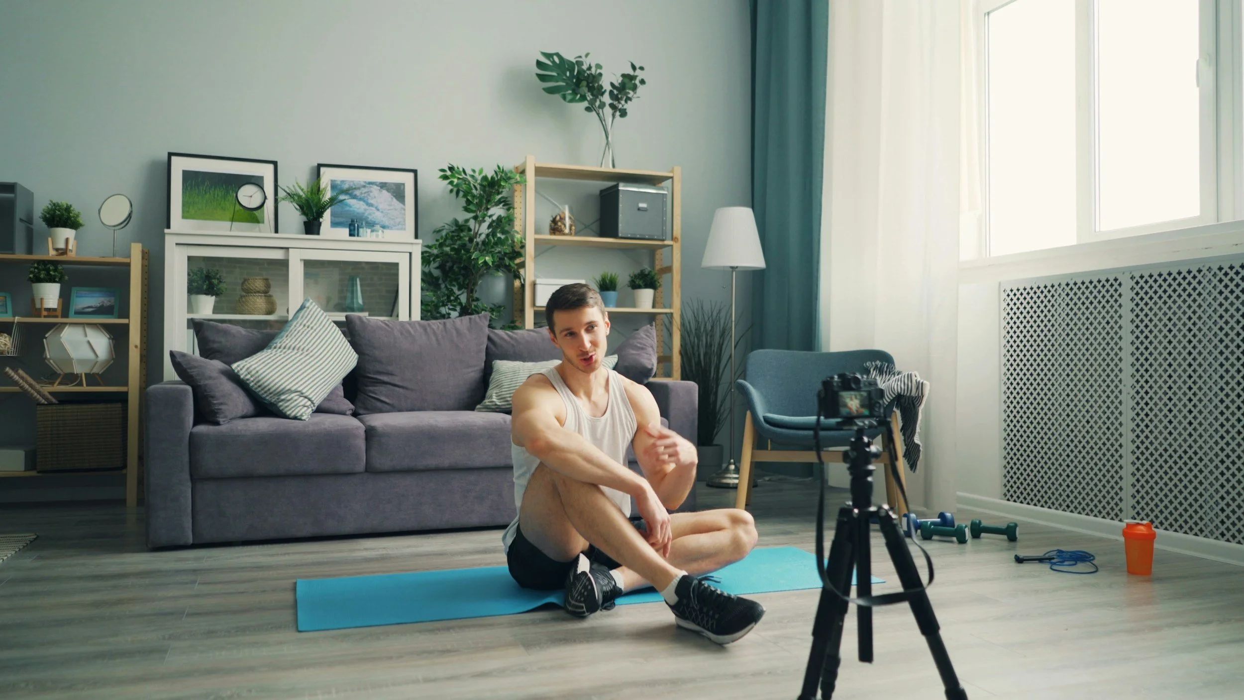 A young man in workout clothes sitting on a yoga mat in a living room, recording himself with a camera on a tripod, with gym equipment and water bottle nearby.