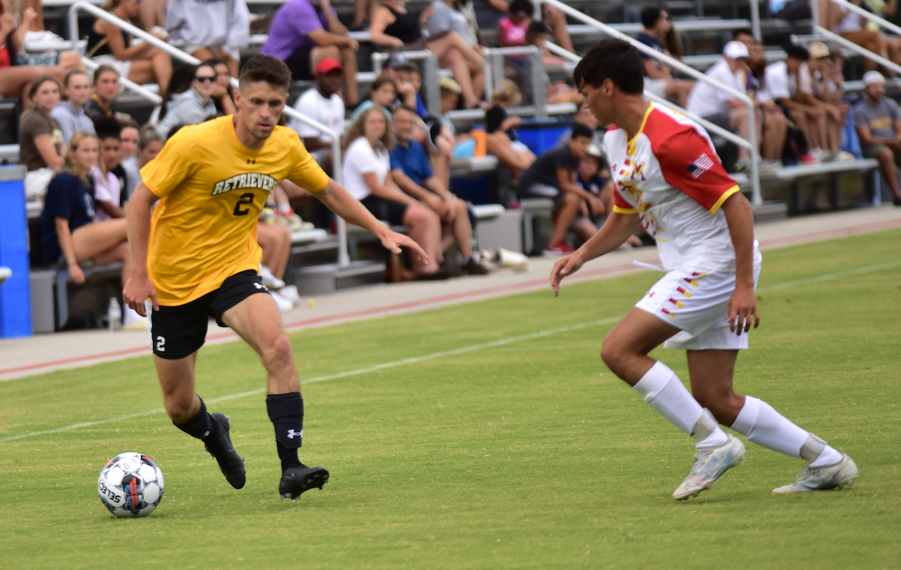 Two soccer players compete for the ball on a field while spectators watch from the stands.
