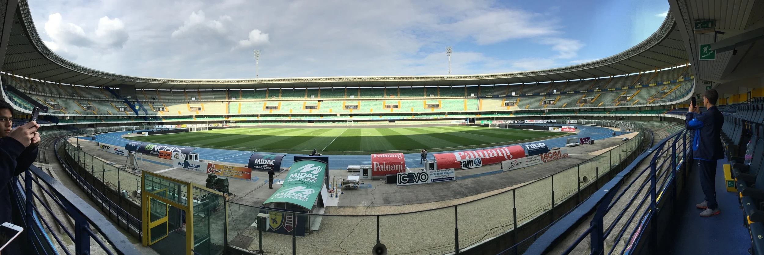 Empty football stadium with a green field, track, and seating tiers, under a partly cloudy sky, with a few spectators taking photos.