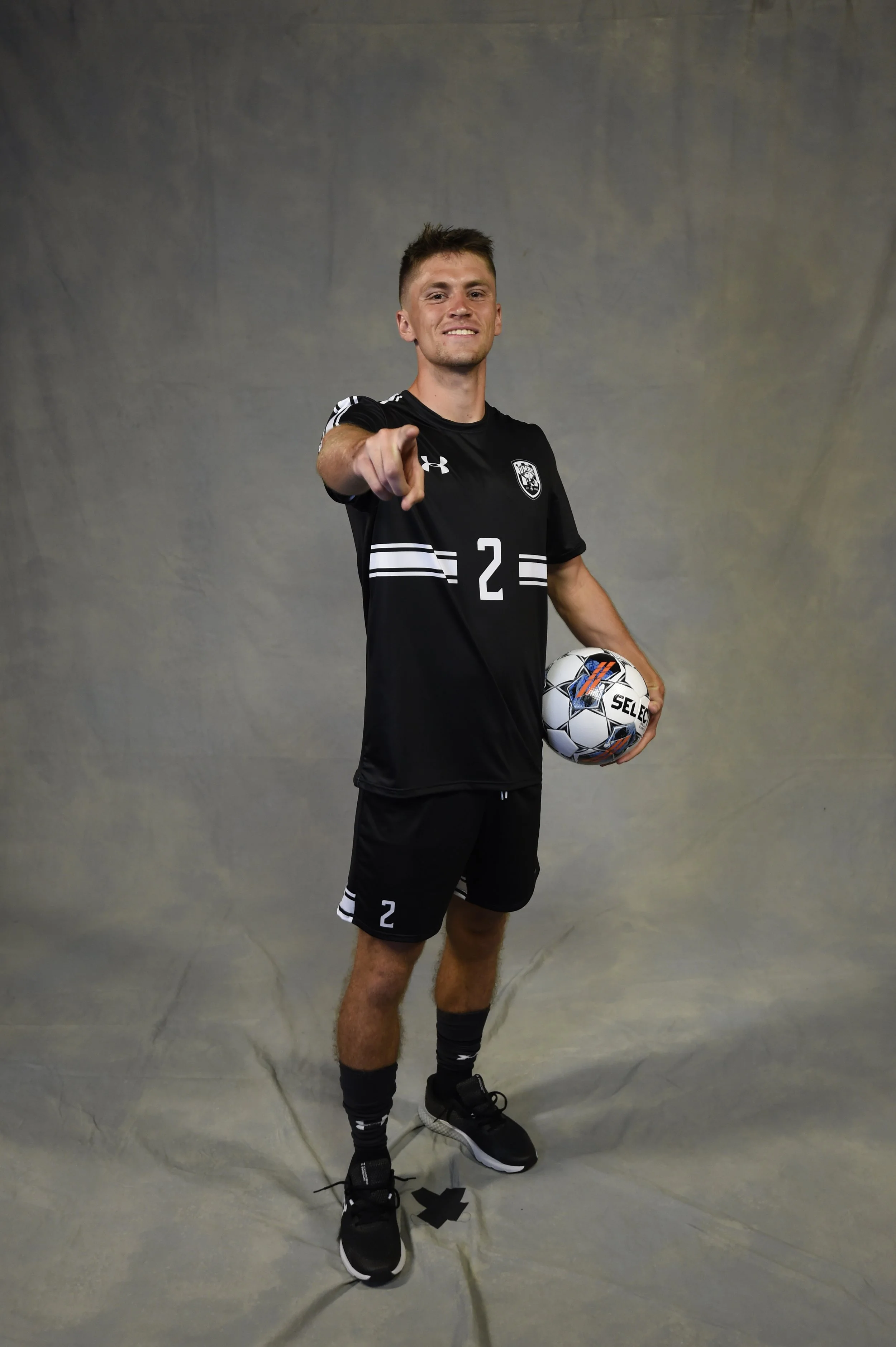 A young man in a black sports uniform with the number 2 on his jersey, holding a soccer ball, and pointing towards the camera in front of a gray background.
