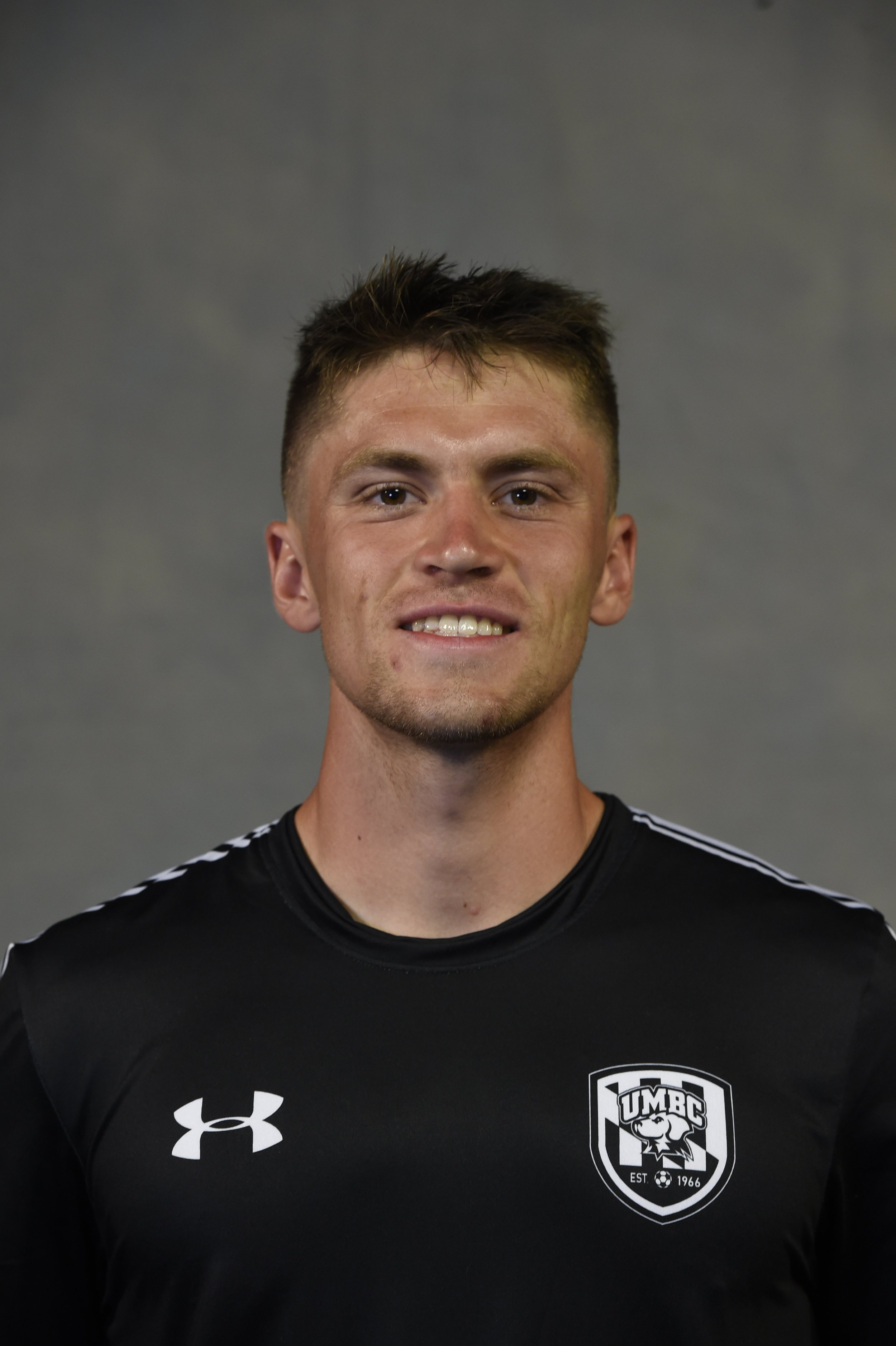 A young man with short brown hair smiling, wearing a black sports jersey with the UMBC logo and lions head emblem on it, standing against a plain gray background.