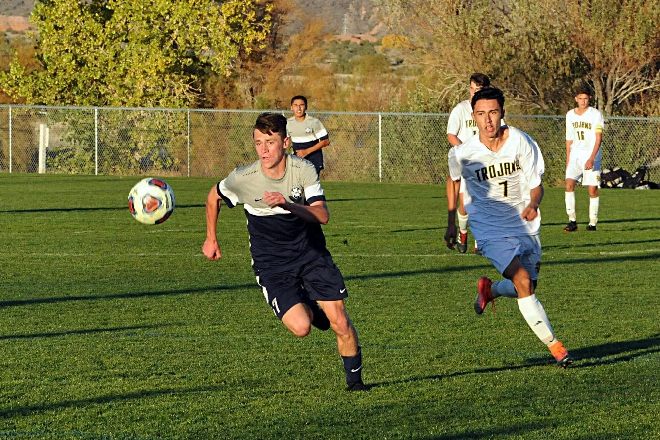 Soccer players in action during a game on a grassy field surrounded by a chain-link fence, with trees and hills in the background.