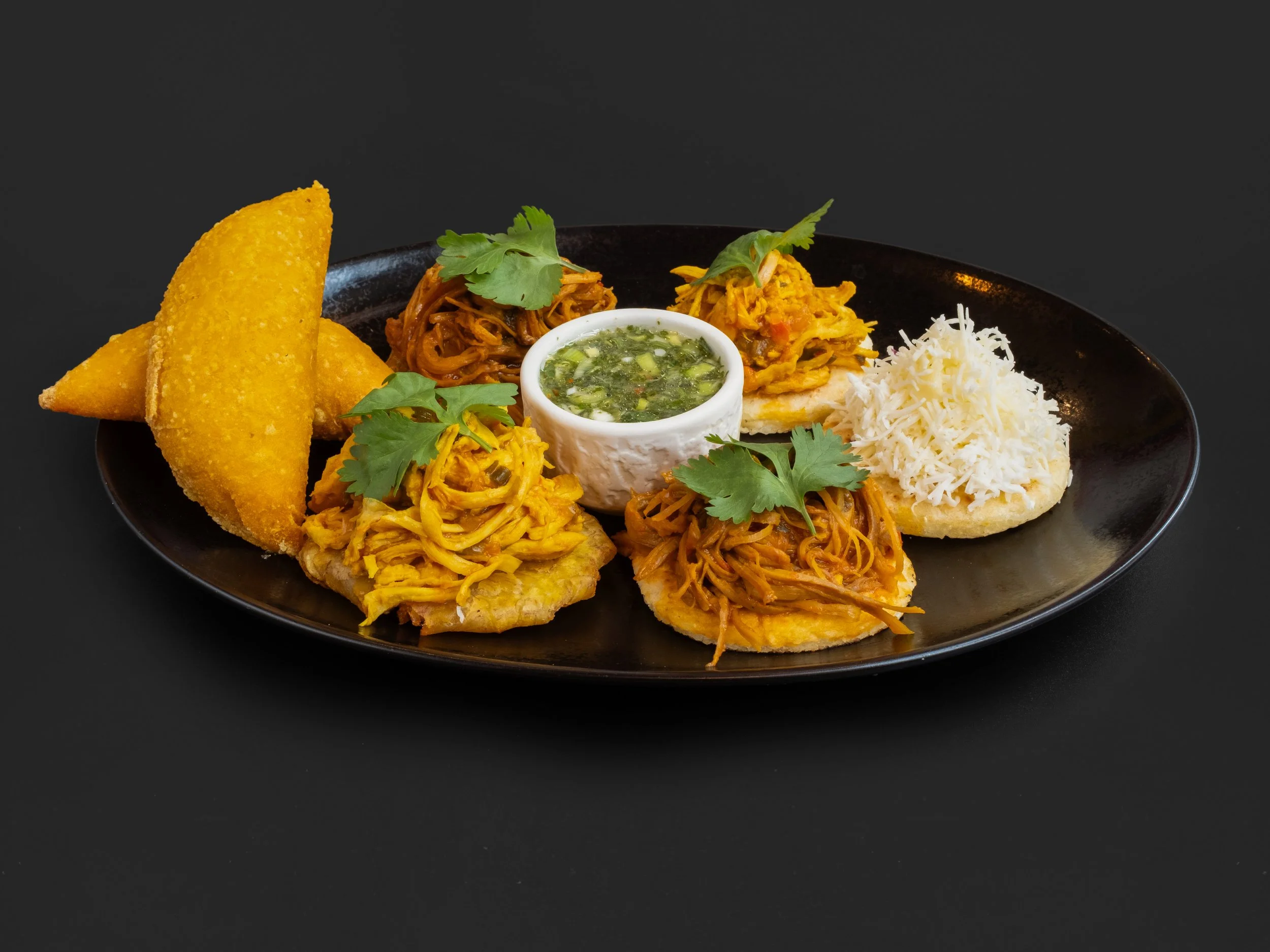A black plate with different types of Indian snacks and side dishes, including golden samosas, shredded chicken, chutney, shredded cheese, and cilantro.
