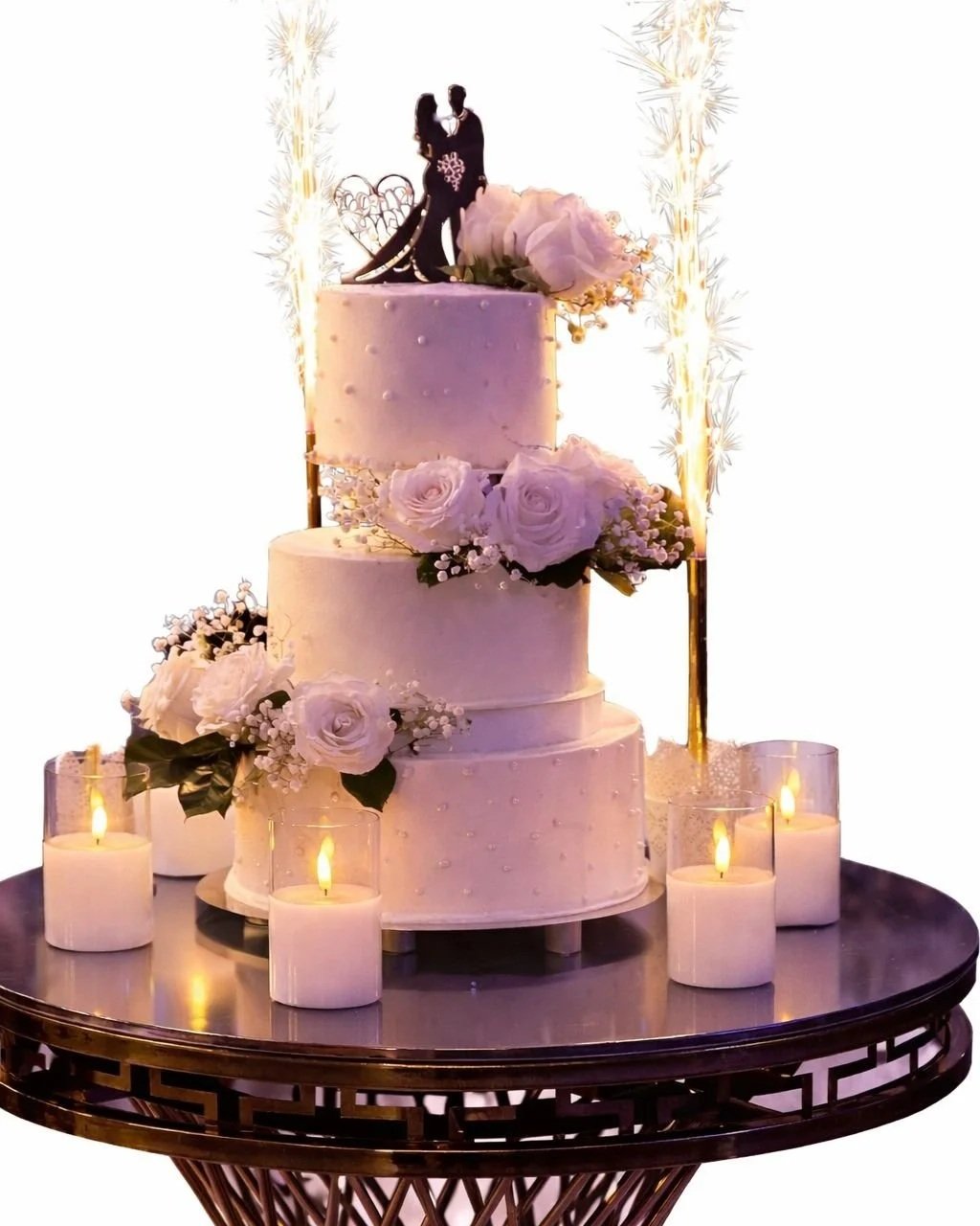 A three-tier wedding cake decorated with white roses and flowers, topped with a silhouette cake topper of a couple, surrounded by candles on a dark table.