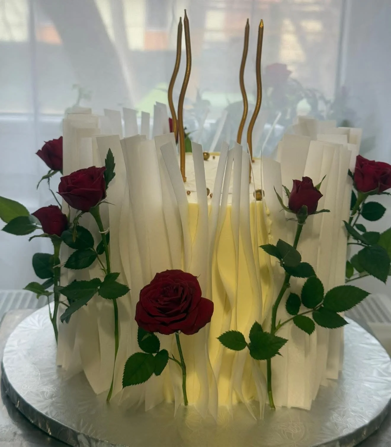A round cake decorated with white chocolate panels, red roses, green leaves, and gold candle-like decorations on top.