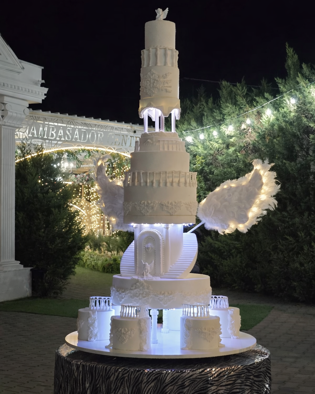 A multi-tiered white wedding cake with angel wings and decorative patterns, displayed on a round table at night, with ➤ lights and greenery in the background.