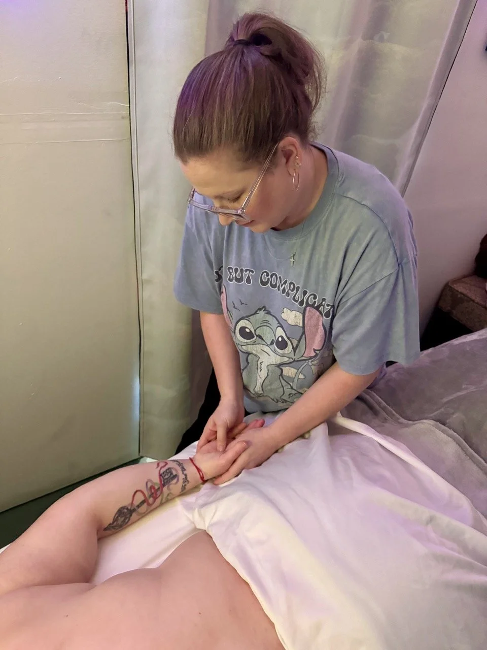 A woman giving a hand massage to a person lying on a massage table. The woman is wearing glasses and a T-shirt featuring Baby Yoda.