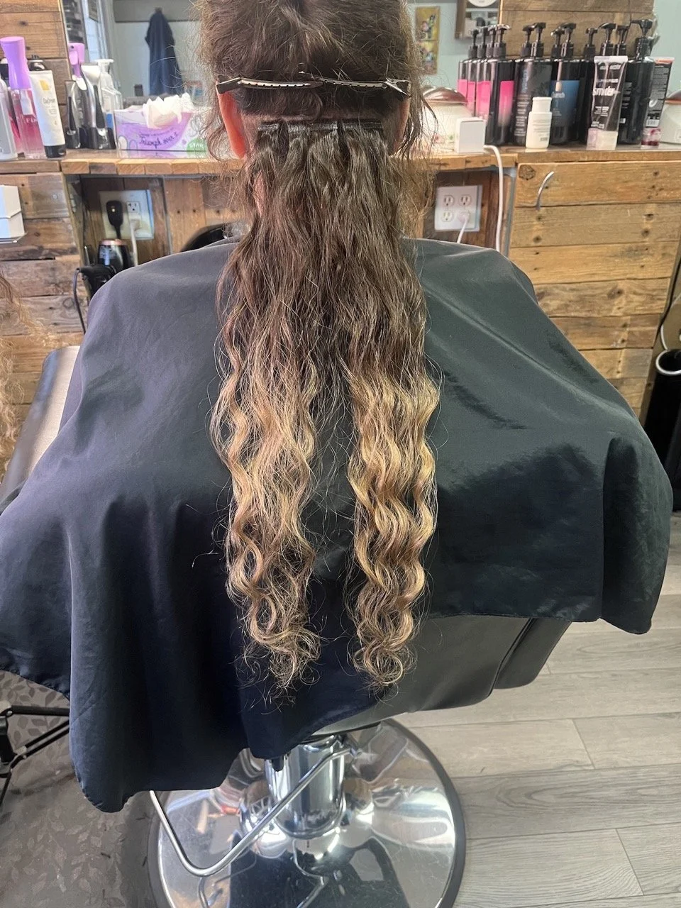Back view of a woman at a hair salon, with long, wavy ombre hair from dark brown to blonde, sitting in a black salon chair over a shiny silver base. The background shows shelves with hair products and a wooden wall.