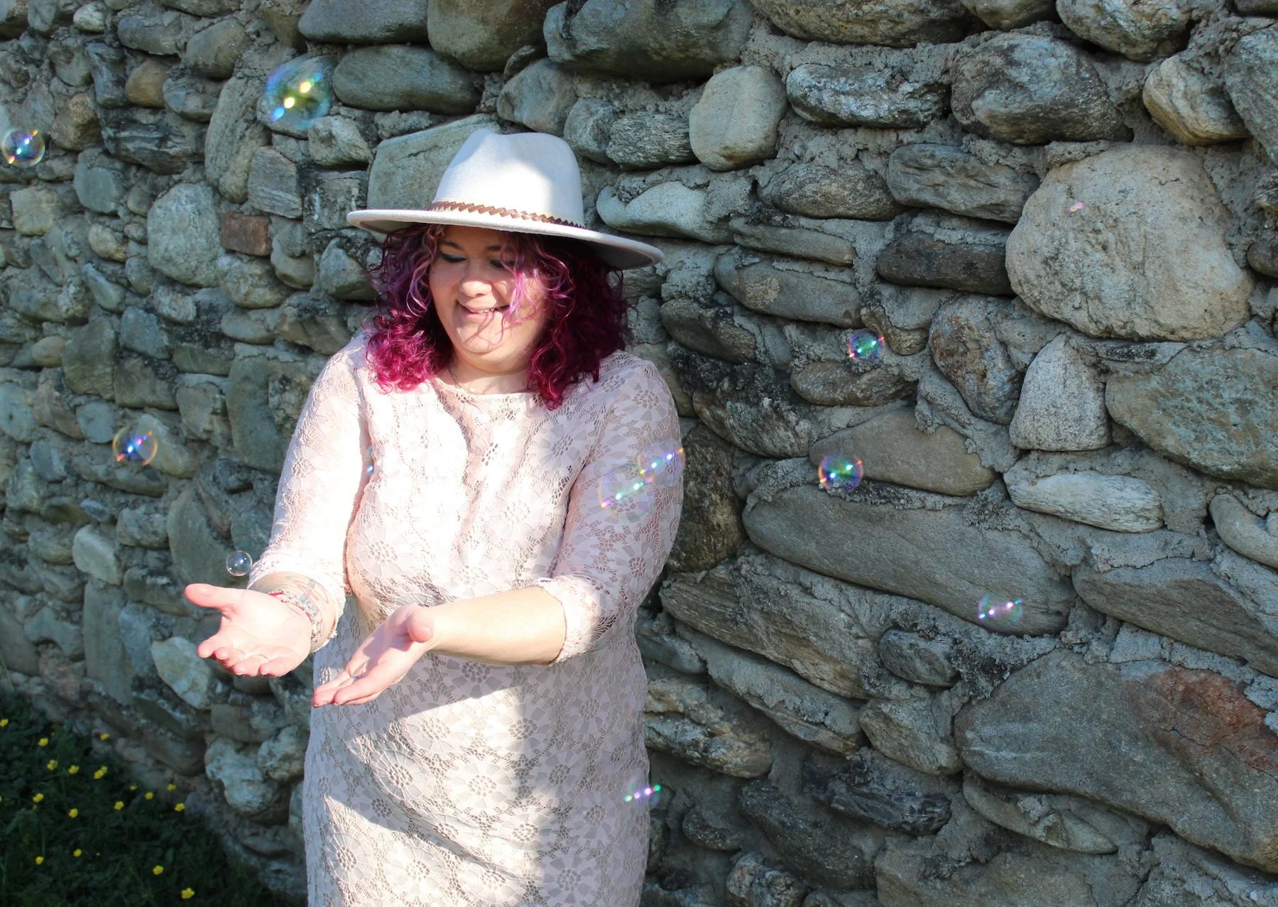 A woman with purple hair and a wide-brimmed white hat stands in front of a stone wall, reaching out to catch floating soap bubbles.