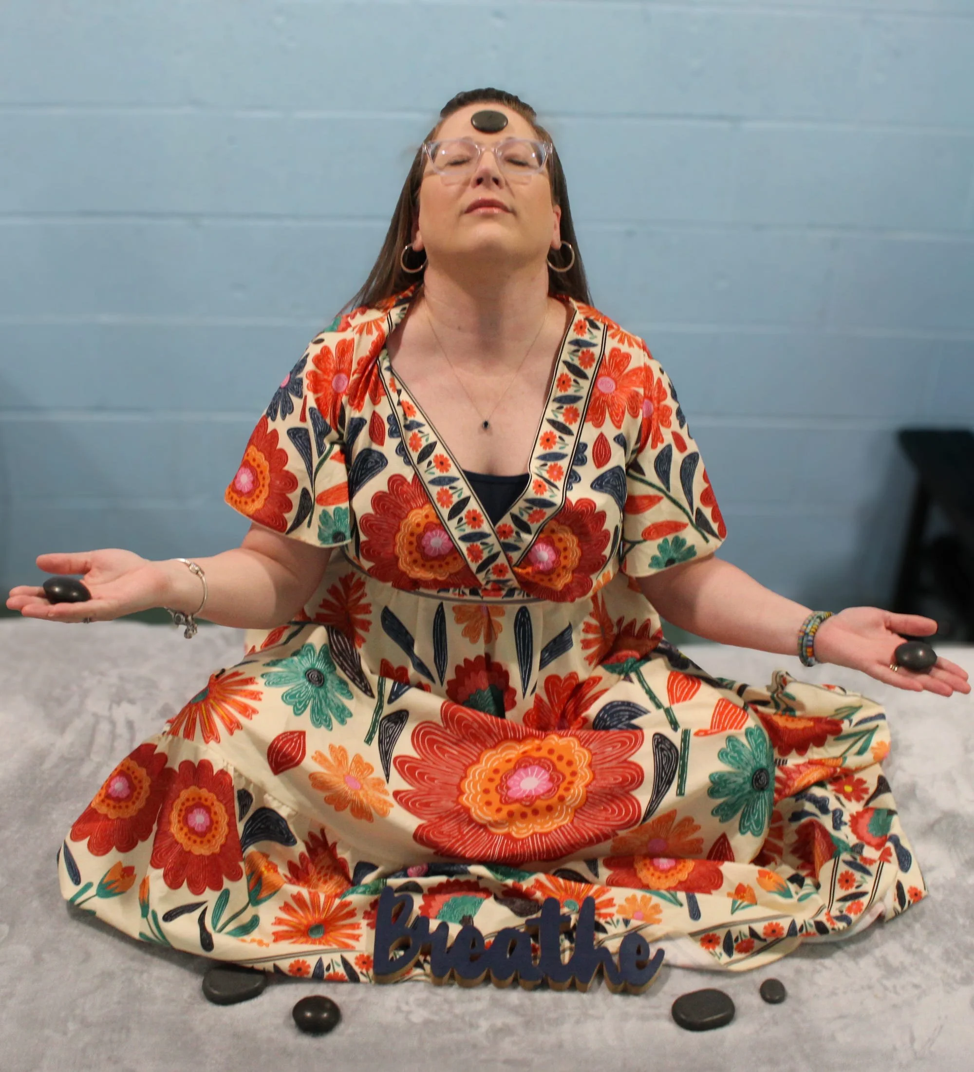 Woman with long dark hair, wearing glasses and earrings, in a colorful floral dress, sitting cross-legged on the floor with her eyes closed and hands resting on her knees, in front of a blue wall.