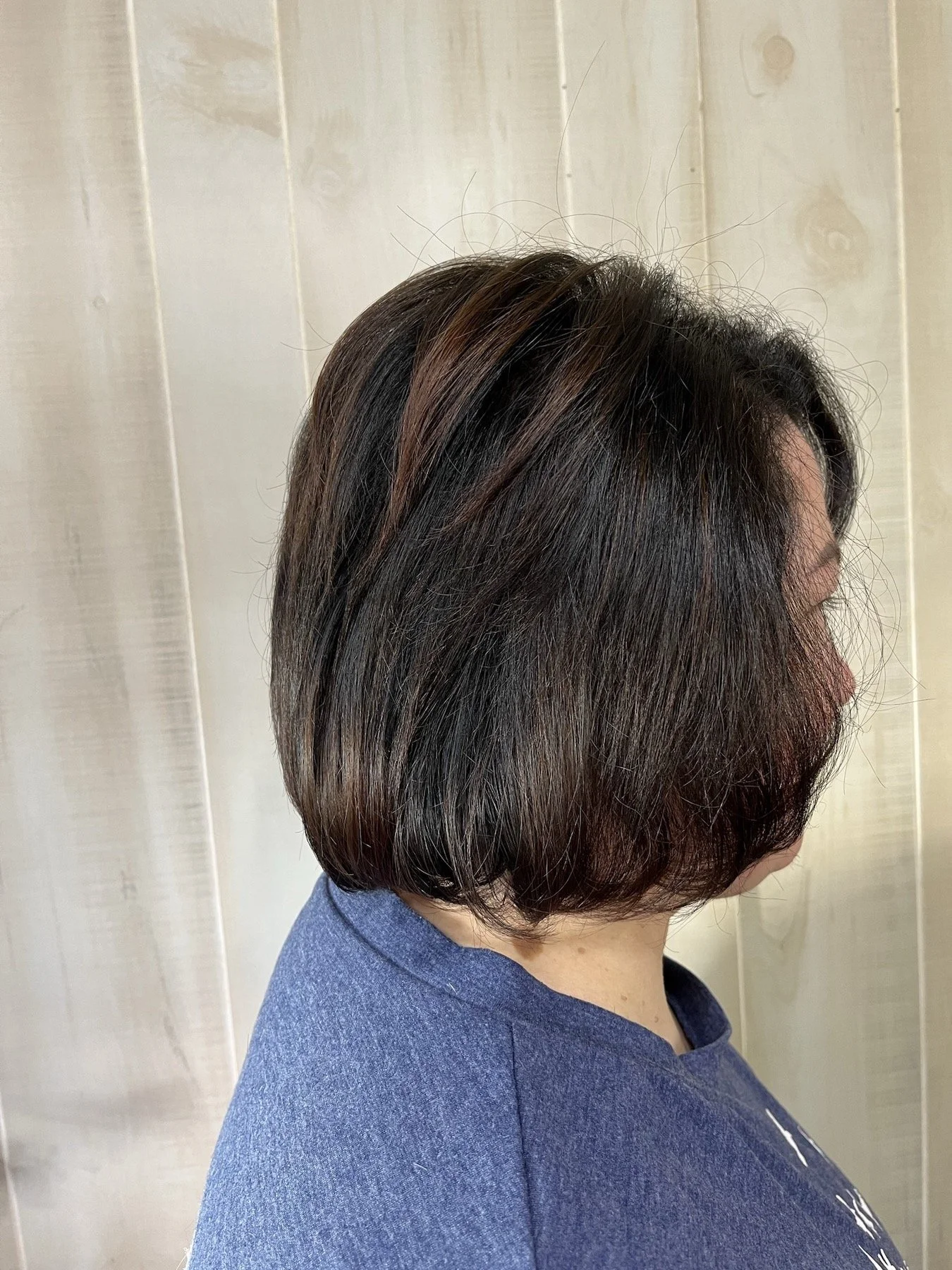 Side view of a woman with short, layered brown hair with subtle highlights, wearing a blue top, standing against a light-colored wooden panel background.