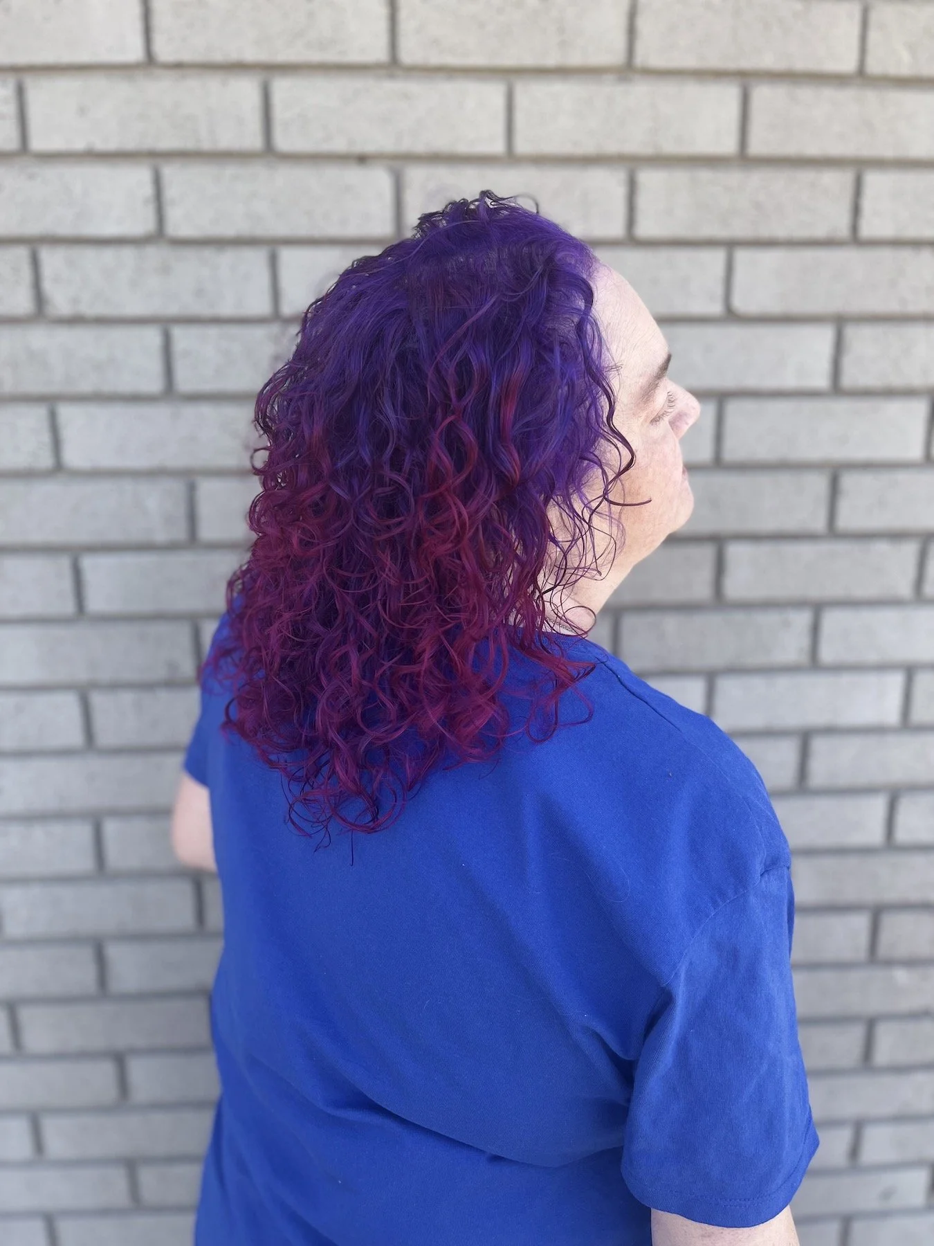 Side view of a person with vibrant purple and pink curly hair, wearing a blue shirt, standing against a brick wall.