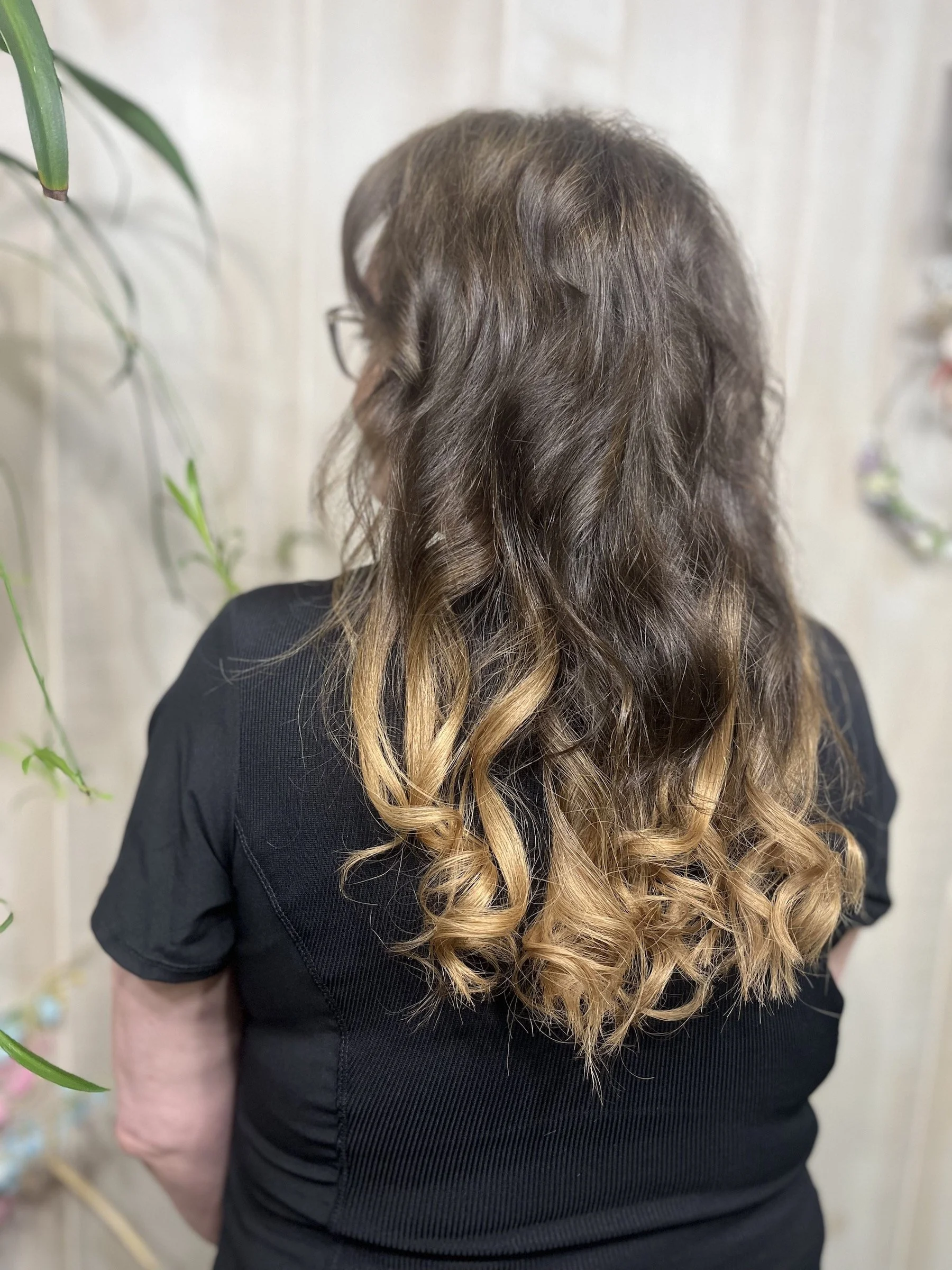 Back view of a woman with long, curly brown hair with blonde highlights, wearing a black top, standing indoors.