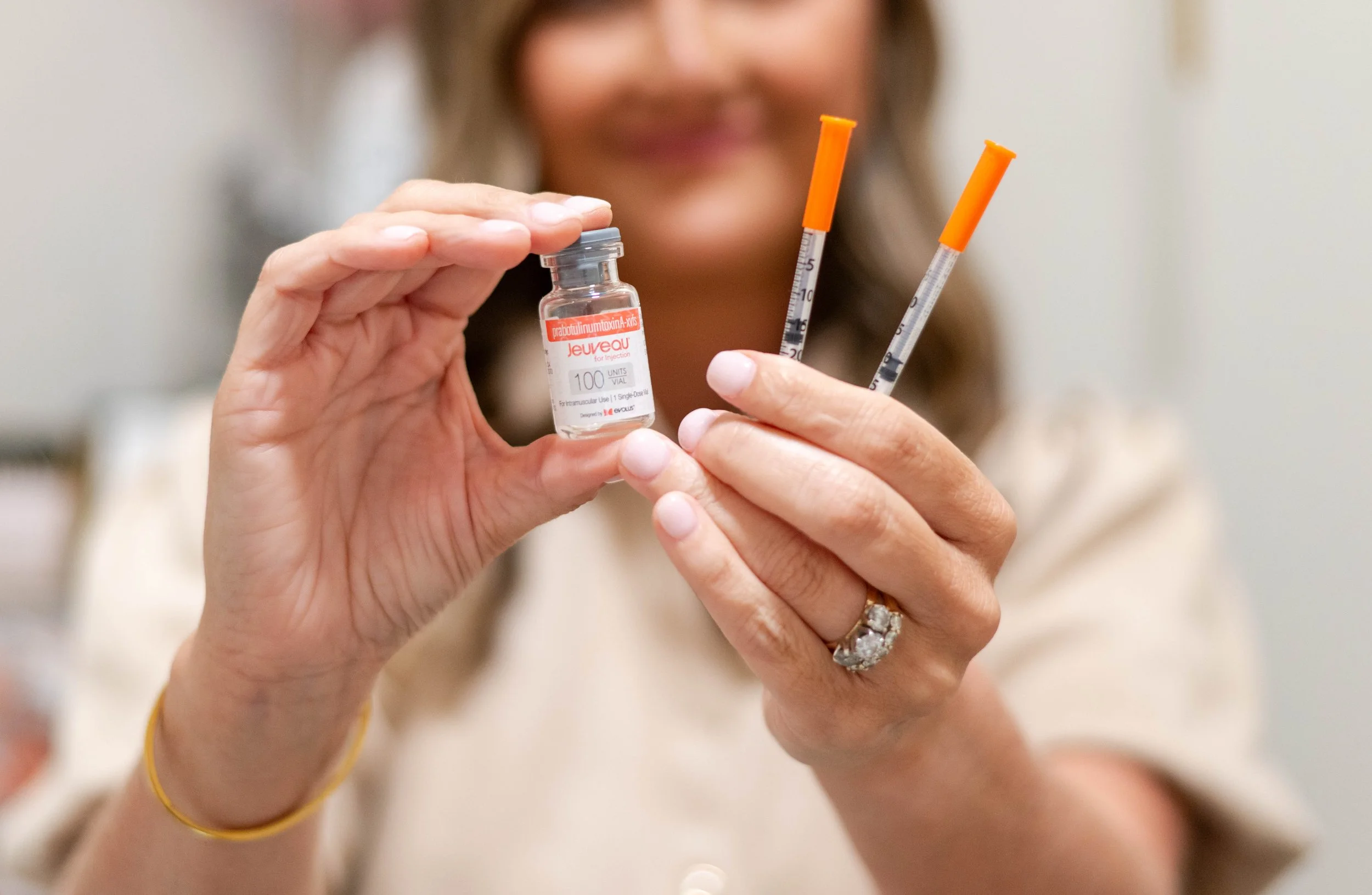 Person holding a small vial labeled 'prabotulinumtoxinA-Xiivs' and two syringes with orange caps, with a blurred woman smiling in the background. Skin Ritual Aesthetics & Wellness in Ridgeville, SC offers Botox, dermal fillers, microneedling, peels
