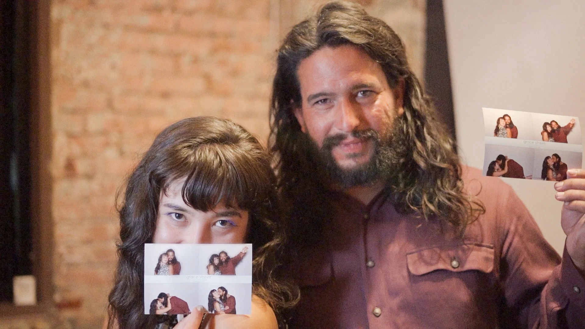A man and a girl holding photo booth strips showing a couple in different poses, with the man having long curly hair and a beard, and the girl having long, straight dark hair with bangs.