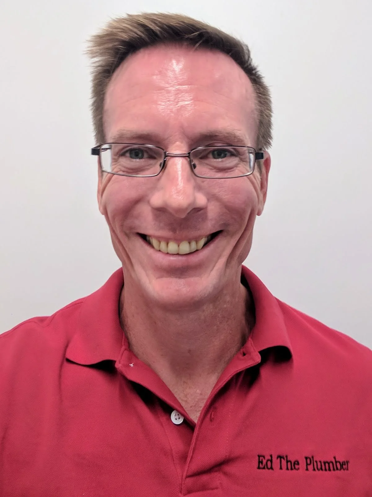A photo of Ed Melancon with a red shirt with the text 'Ed The Plumber' embroidered on it.