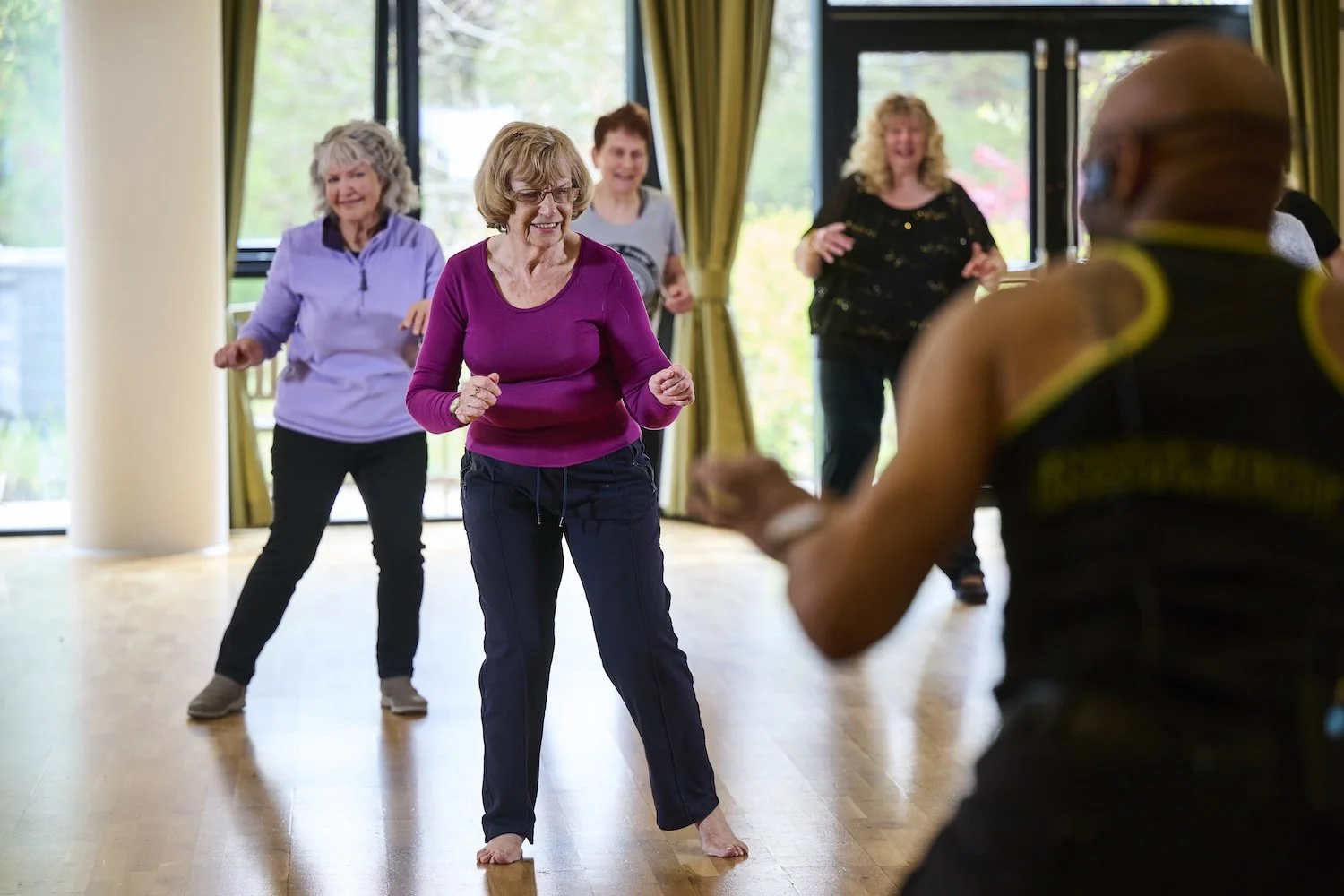 Older women dancing in a room with large windows, smiling and having fun, with an instructor leading the activity.