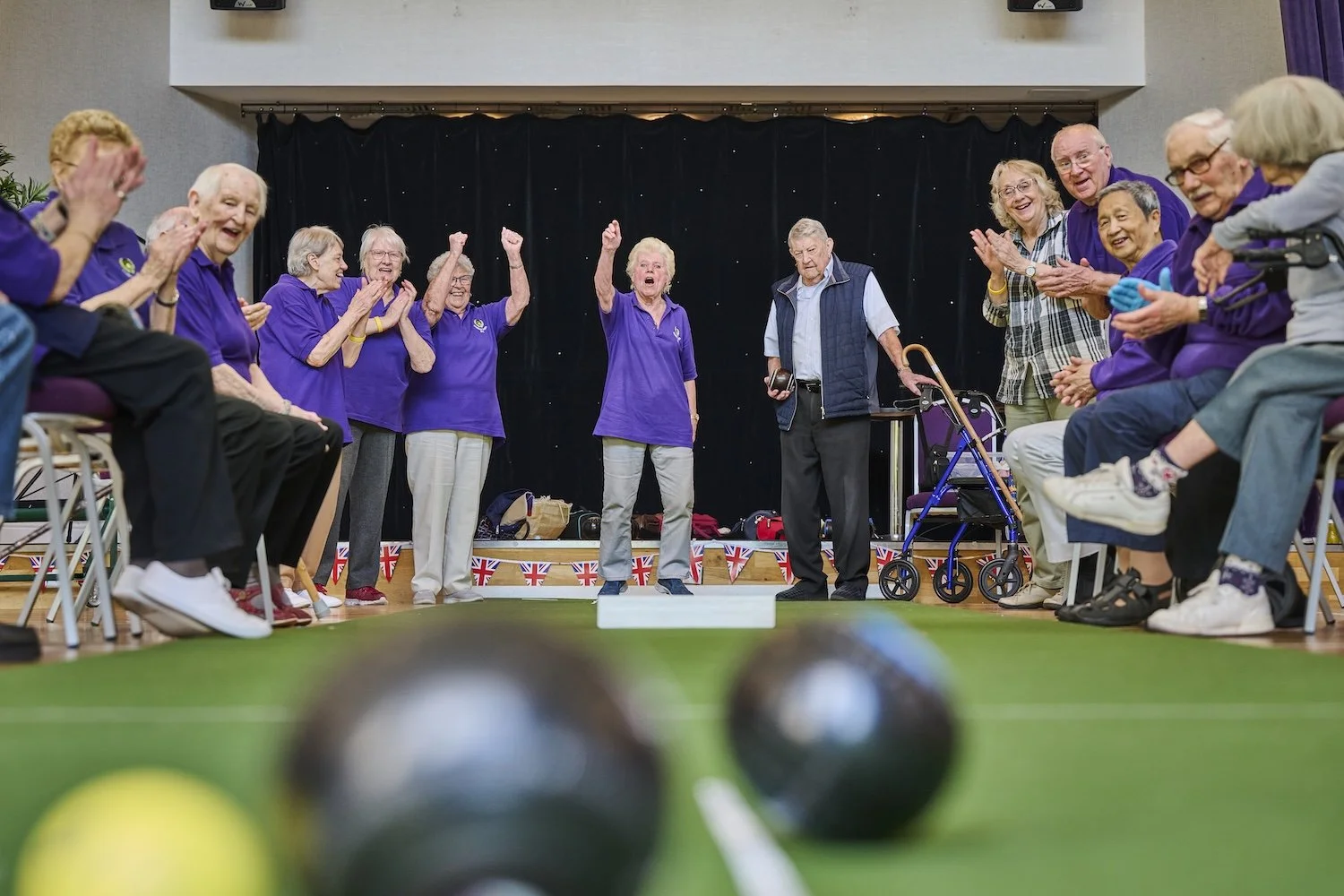 Elderly people playing bocce ball indoors, some standing and cheering, others sitting and watching, with a black curtain behind them and Union Jack bunting along the stage edge.