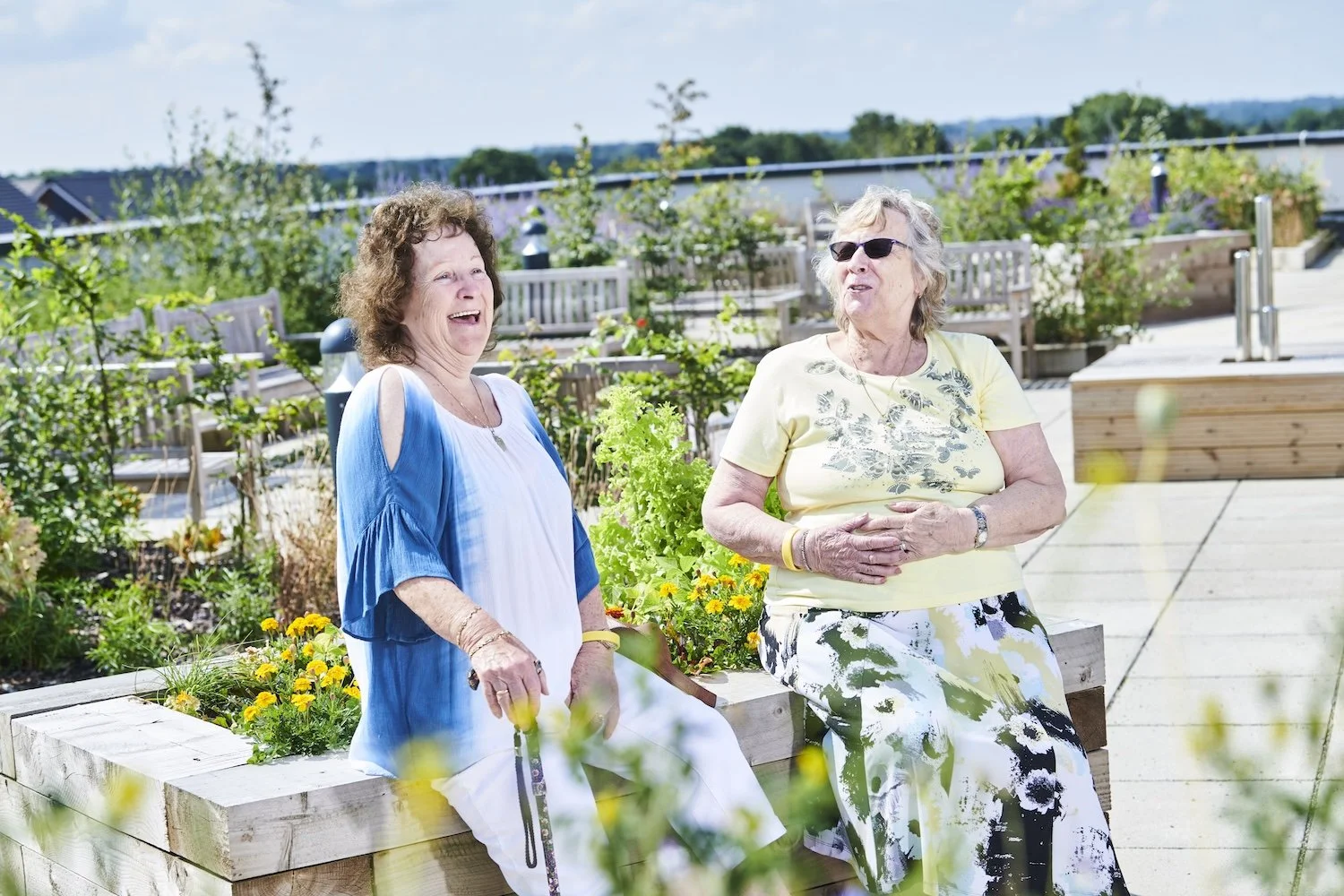 Two elderly women sitting on a wooden bench in an outdoor garden area, smiling and engaging in conversation surrounded by plants and flowers with a clear sky in the background.