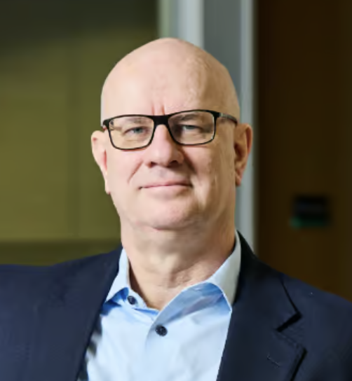 Headshot of a smiling older man with glasses, wearing a suit and light blue shirt, indoors.