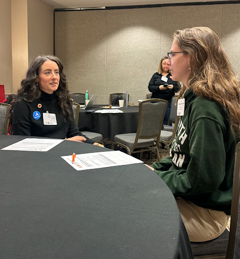 Two women sitting at a round table engaged in conversation at a professional event or conference, with name tags visible on their clothing, and a third woman standing in the background.