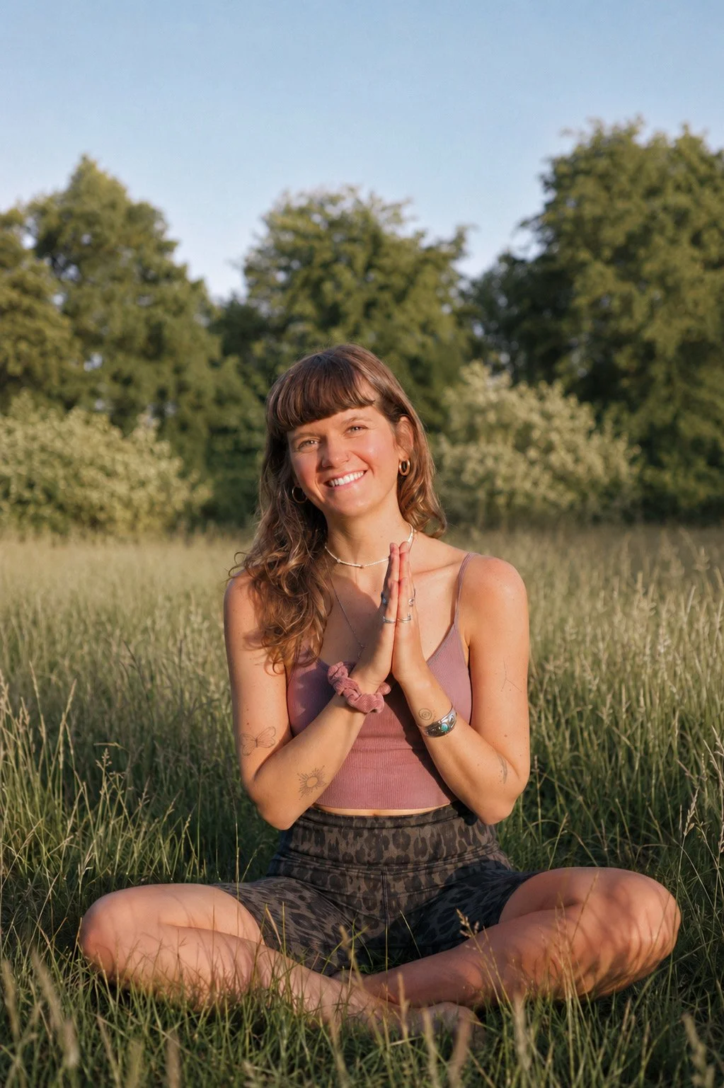 Junge Frau sitzt im Gras in einem Park, lächelnd mit zusammengelegten Händen in Gebet- oder Meditationshaltung, bei Sonnenuntergang.