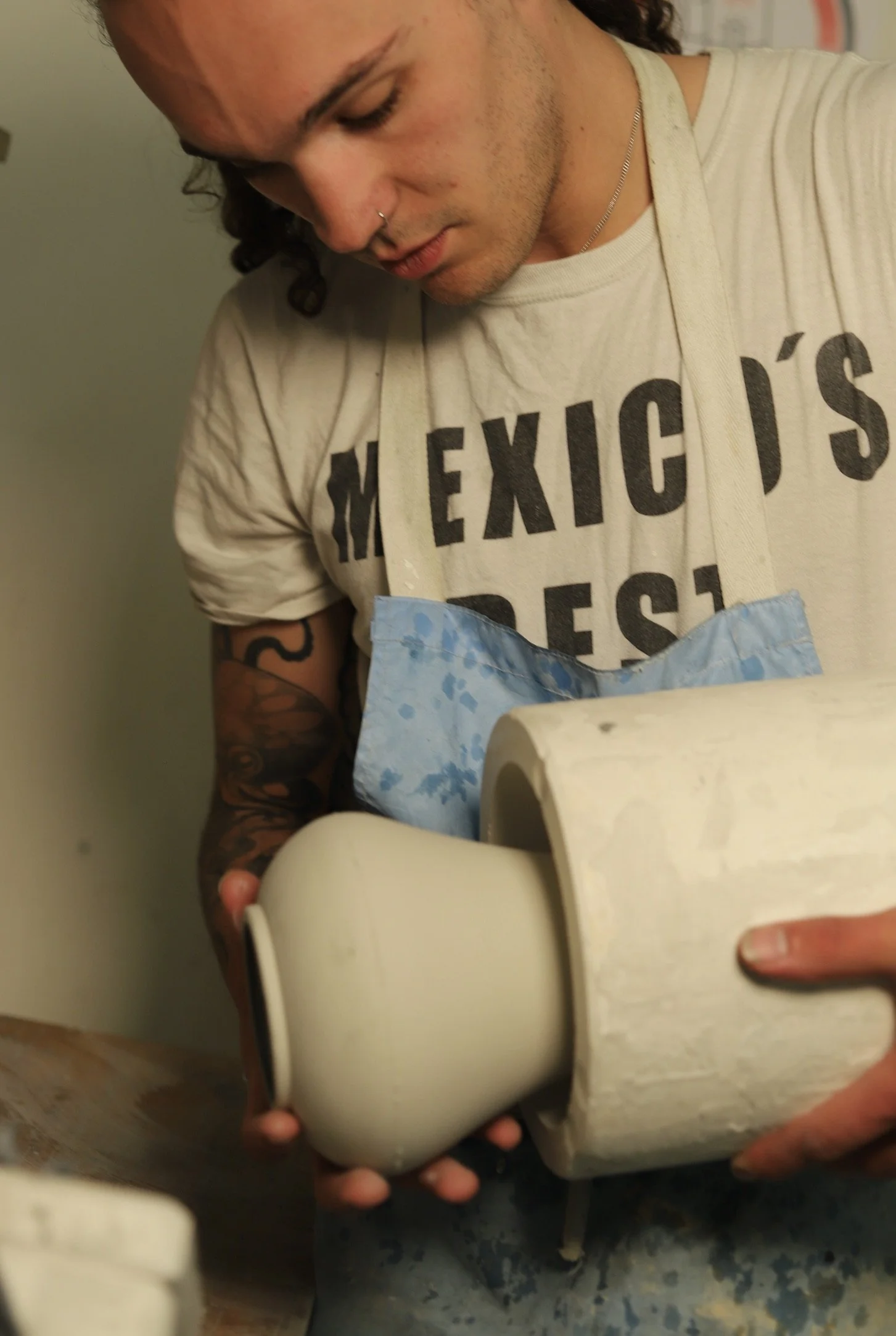 A person with dark hair, a nose piercing, and a tattoo on their arm holds a roll of toilet paper, partially unrolled, while standing in a room with a greenish wall.