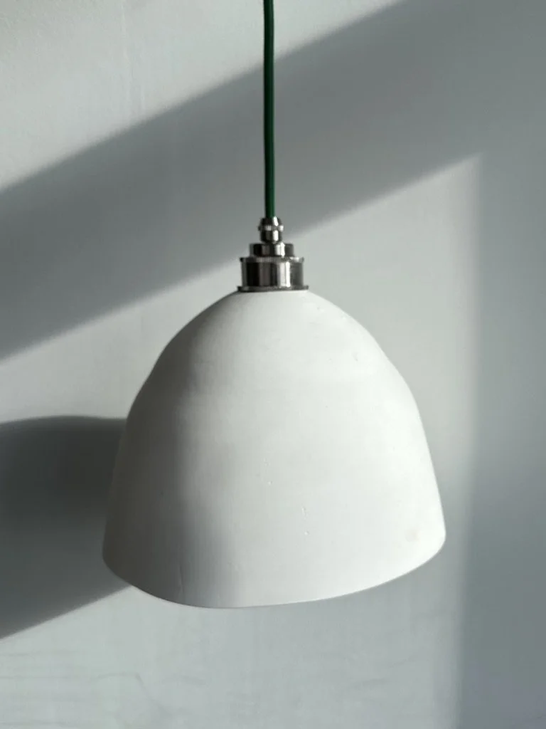 A white hanging pendant lamp with a metal fitting at the top, suspended from a green cord against a plain wall with shadows.