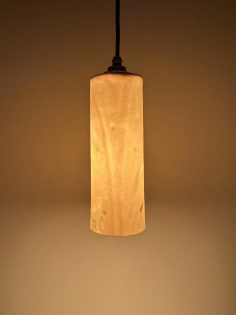 A hanging ceiling lamp with a cylindrical beige lampshade, turned on and emitting a warm light.