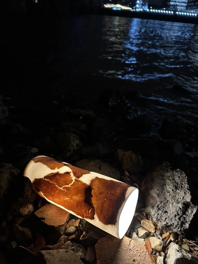 A ceramic mug with a brown and white marbled design lying on rocky ground by a body of water at night. City lights are visible in the distance across the water.