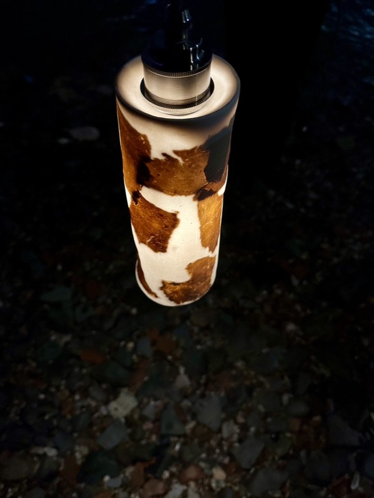 A vape with a cowhide pattern on the body, hanging from a dark background above a rocky surface.