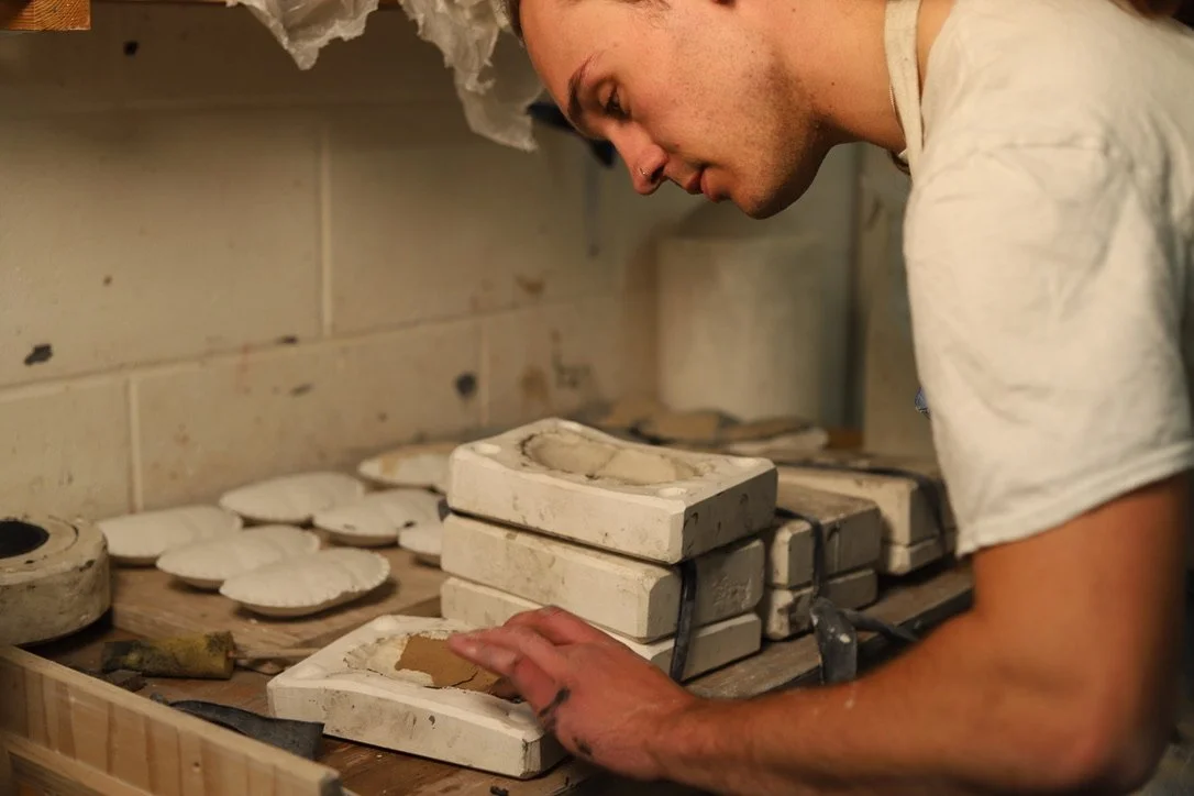 A person working on ceramic molds in a pottery studio, with stacks of molds and pottery tools on the workbench.