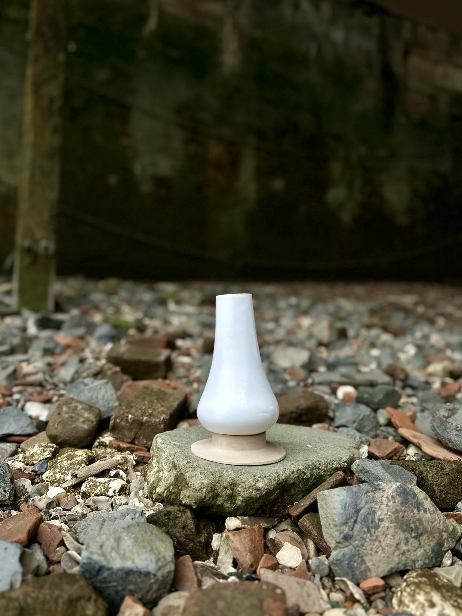 A white object with a rounded base and narrow top sits on a stone in a rocky outdoor area, with a dark, mossy structure in the background.