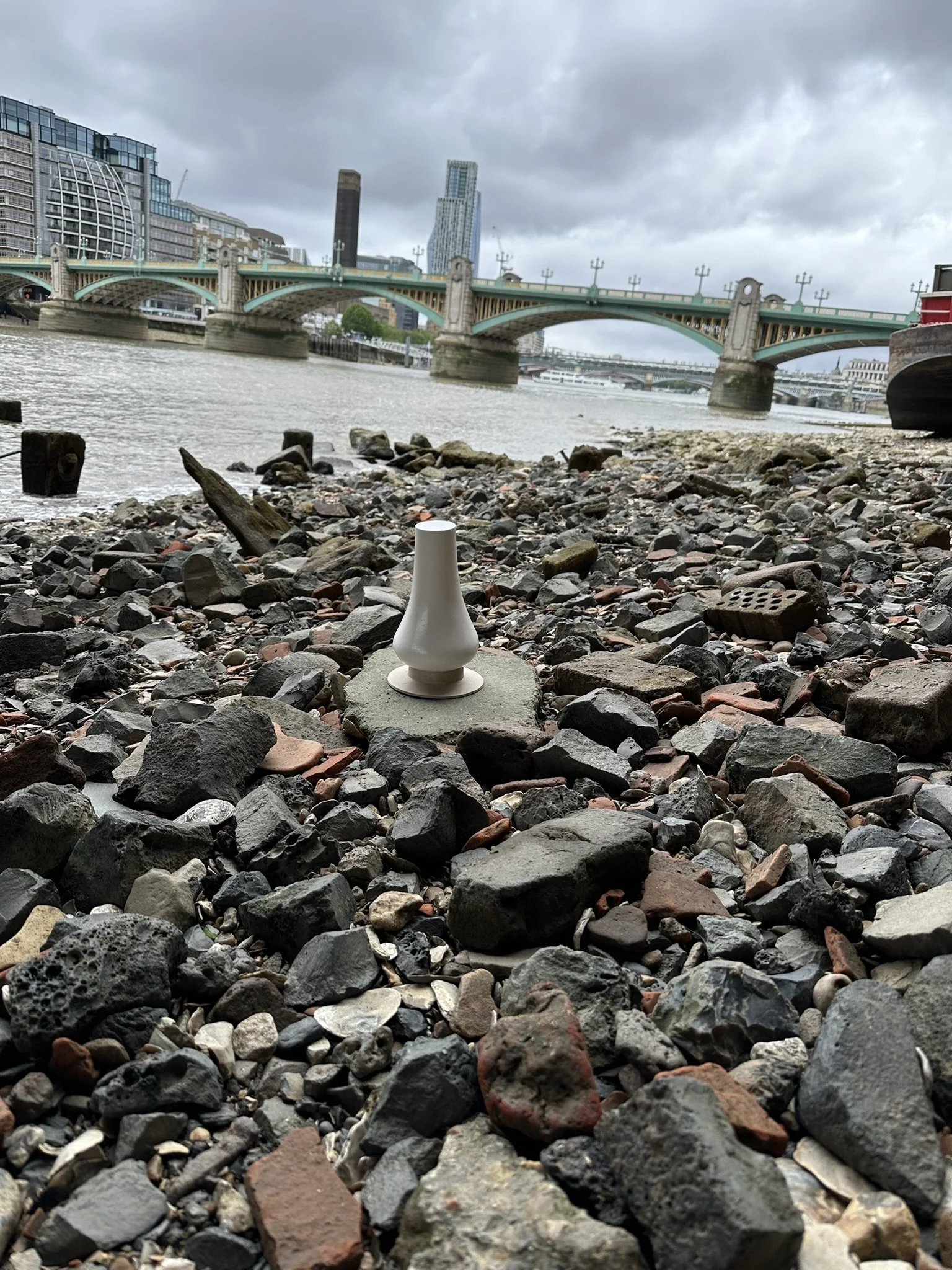 View of a rocky riverbank with a white ceramic vase placed on a large rock in the foreground, a bridge and city skyscrapers under cloudy sky in the background.