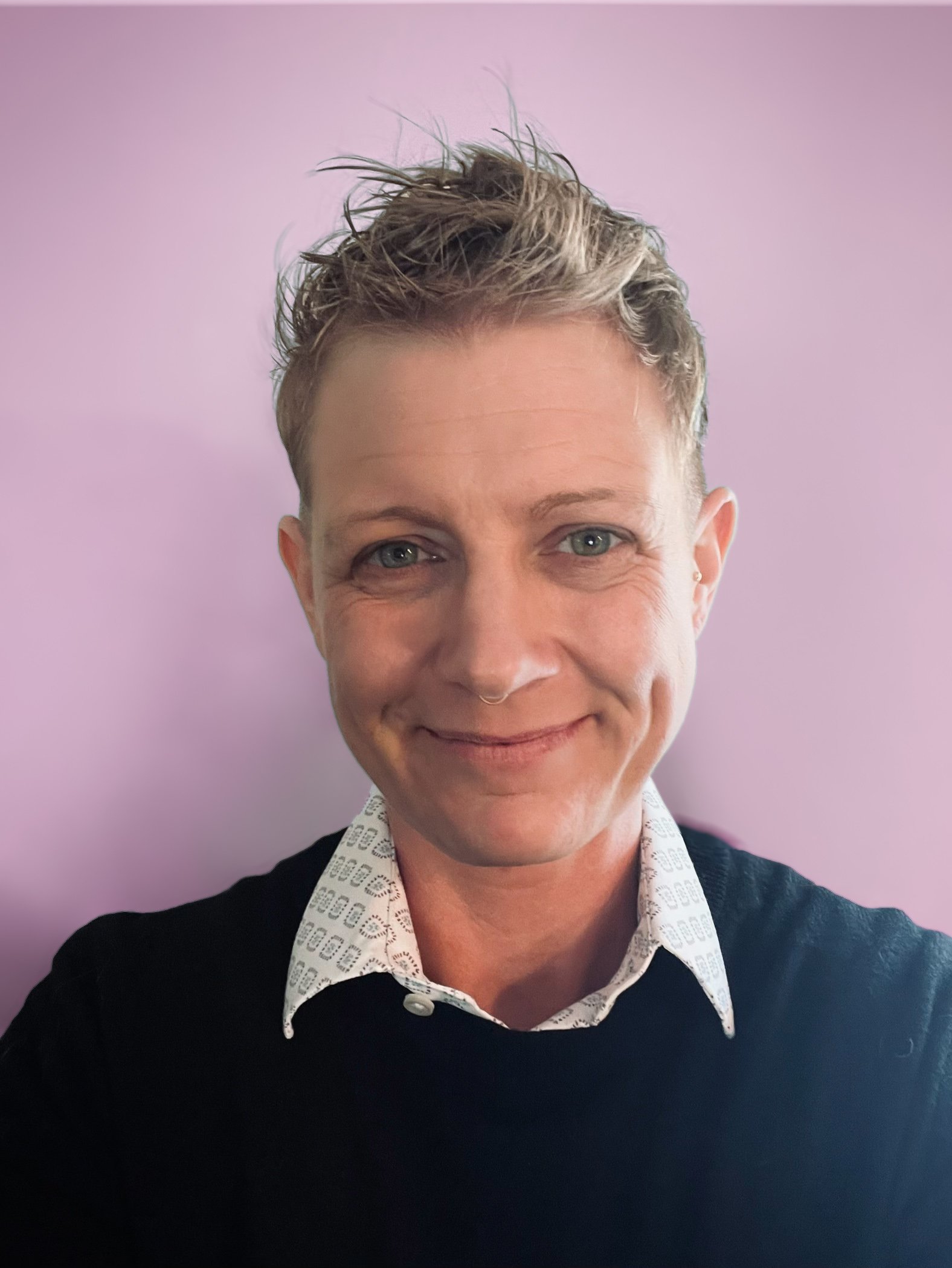 A woman with short blonde hair styled in a spiky manner, smiling, wearing a black sweater over a white collared shirt with a geometric pattern, standing against a light purple background.