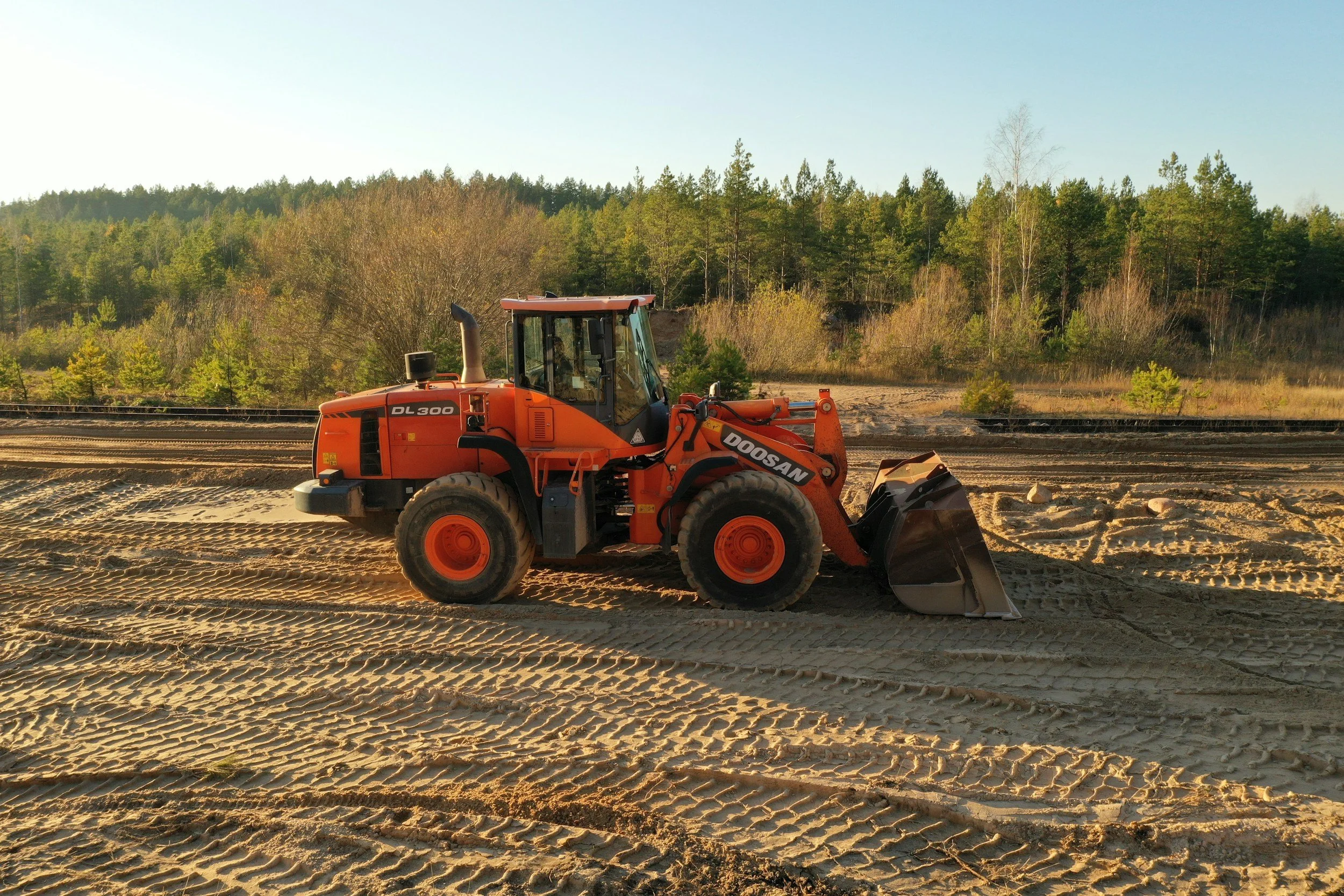 A Doosan DL300 wheel loader working on a construction site with sand and dirt, with railroad tracks and trees in the background during daytime.