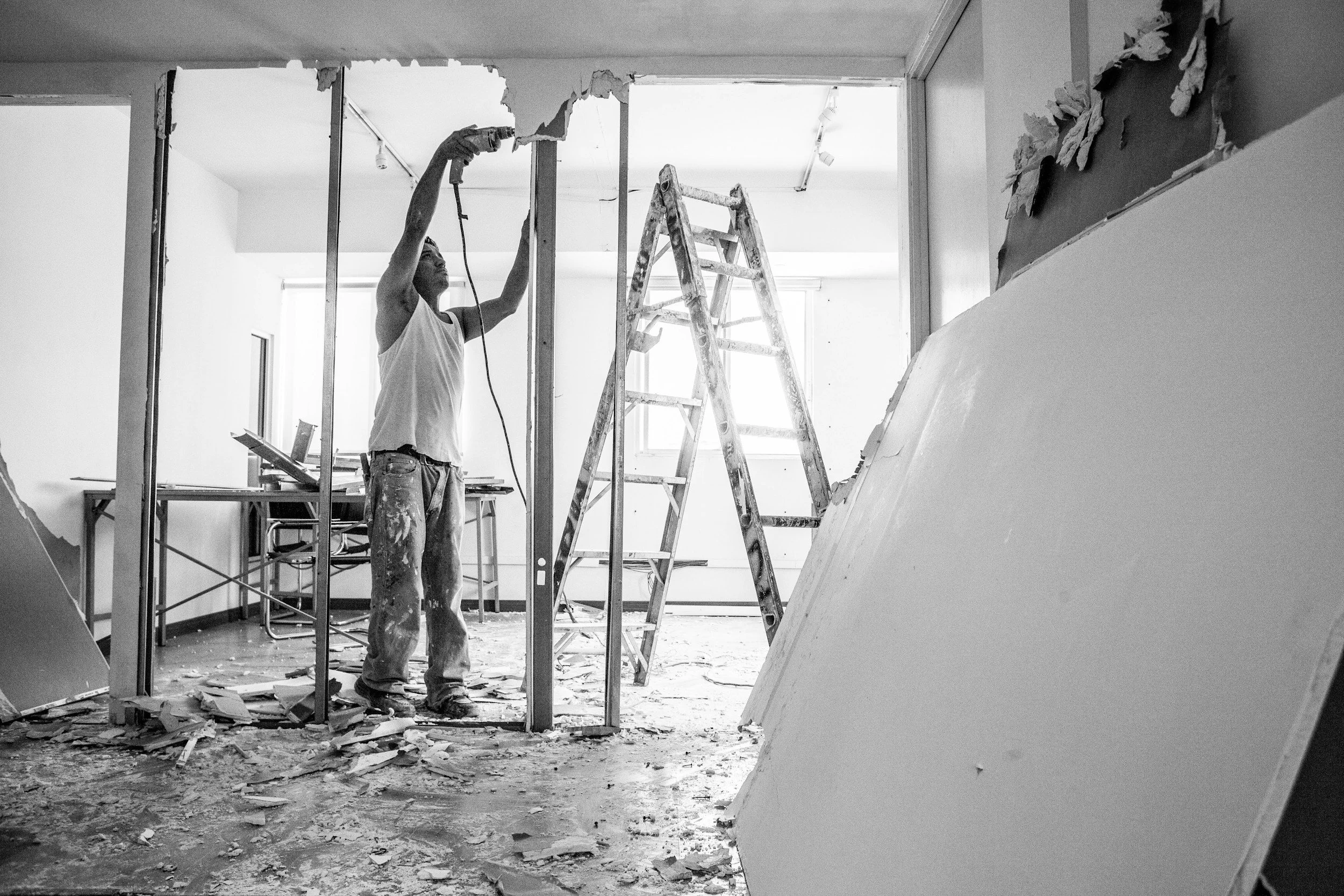 A man is working on tearing down a wall in a room under renovation, using a power tool. Debris is scattered on the floor, and a ladder and furniture are visible in the background.