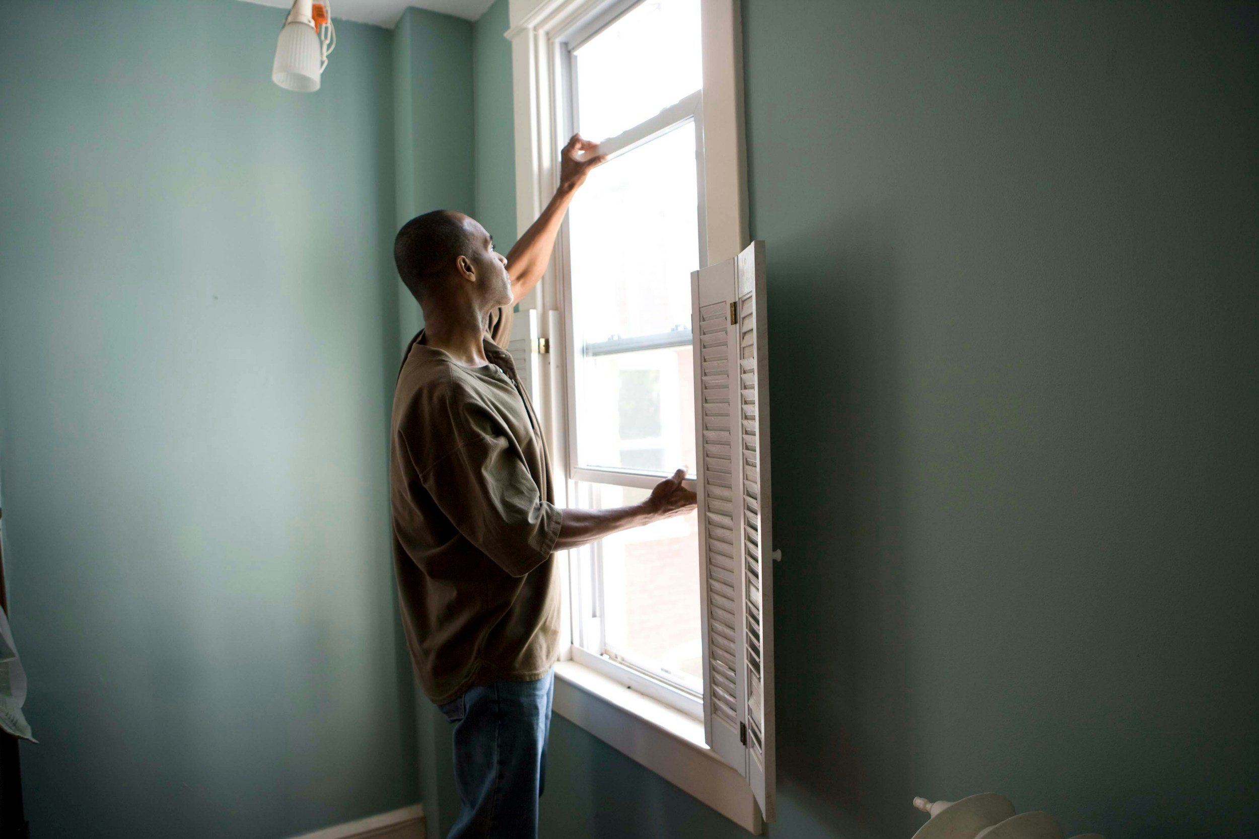 A man opening a window in a room with turquoise walls, a wooden window frame, and a white wall-mounted light fixture on the ceiling.