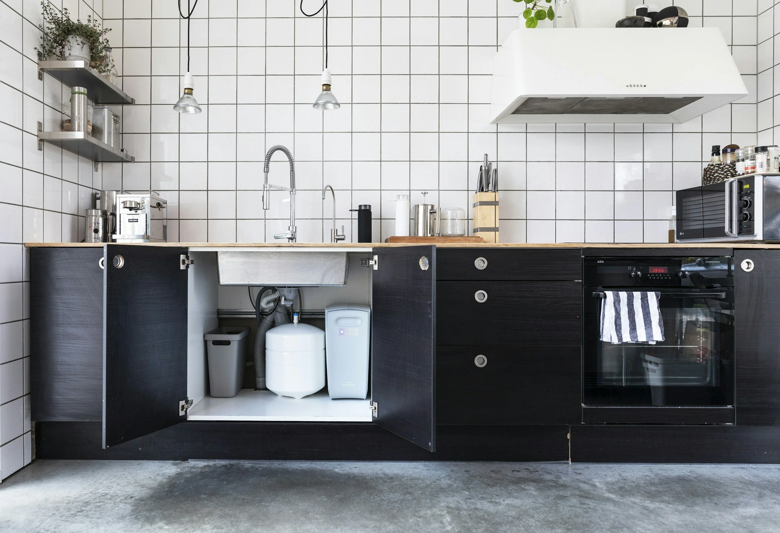 Modern kitchen with black cabinets, white tiled walls, and stainless steel appliances. Open cabinet with a trash bin and water filtration system underneath the sink. Countertop with a knife block, a toaster oven, and various kitchen items. Shelves with jars and containers. Overhead lights hanging above the counter.