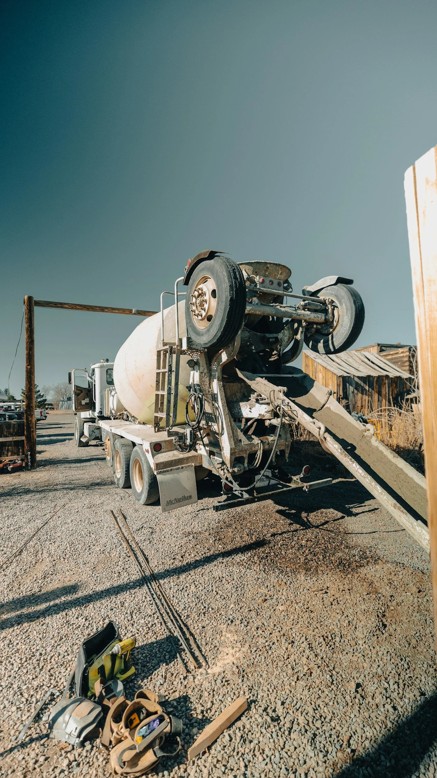 A cement mixer truck designed to look like an overturned car, with tools and construction equipment on the gravel ground nearby, and a clear sky overhead.