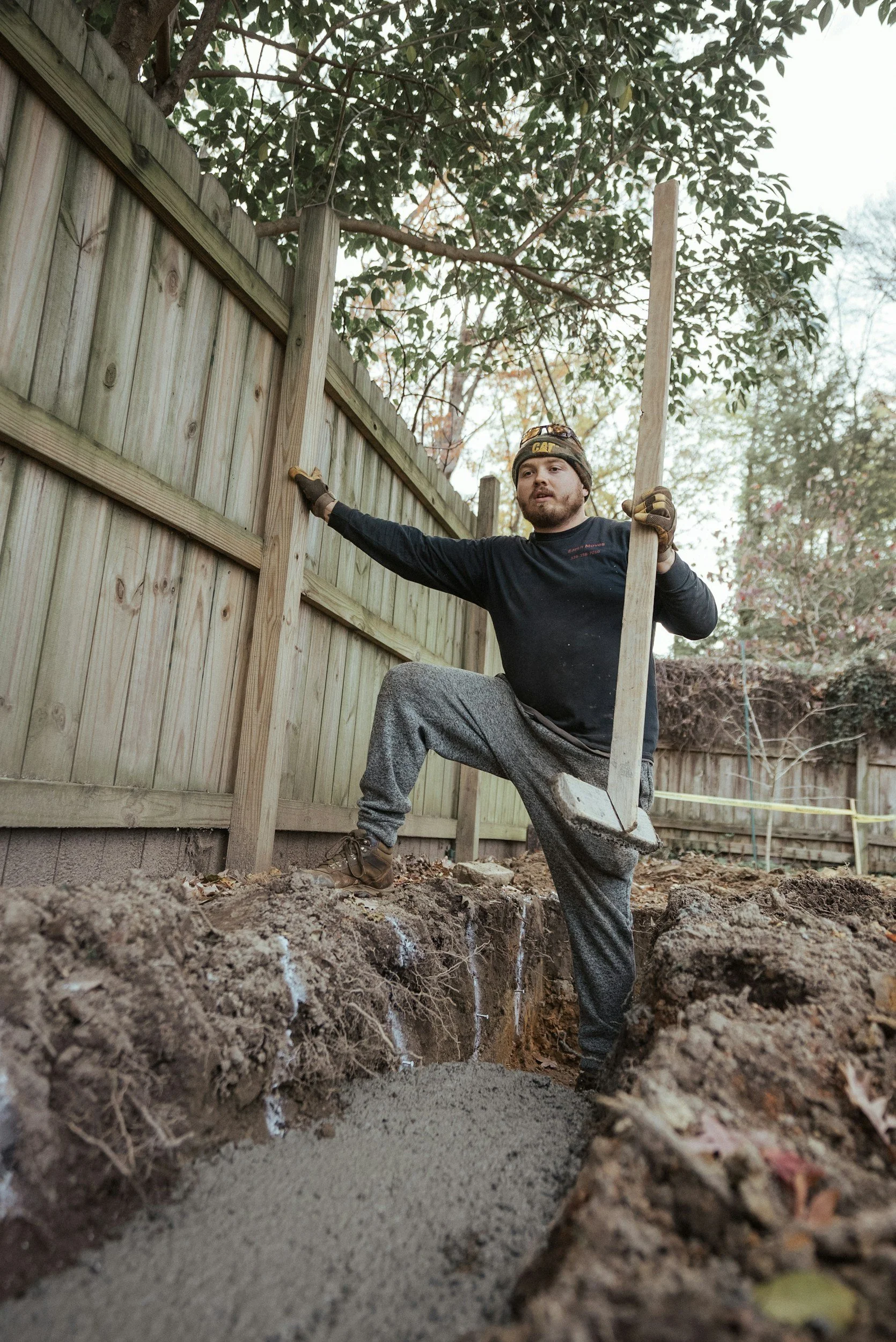 A man working on a home improvement project outdoors, holding a wooden post and standing in a trench filled with dirt and concrete, with a wooden fence and trees in the background.